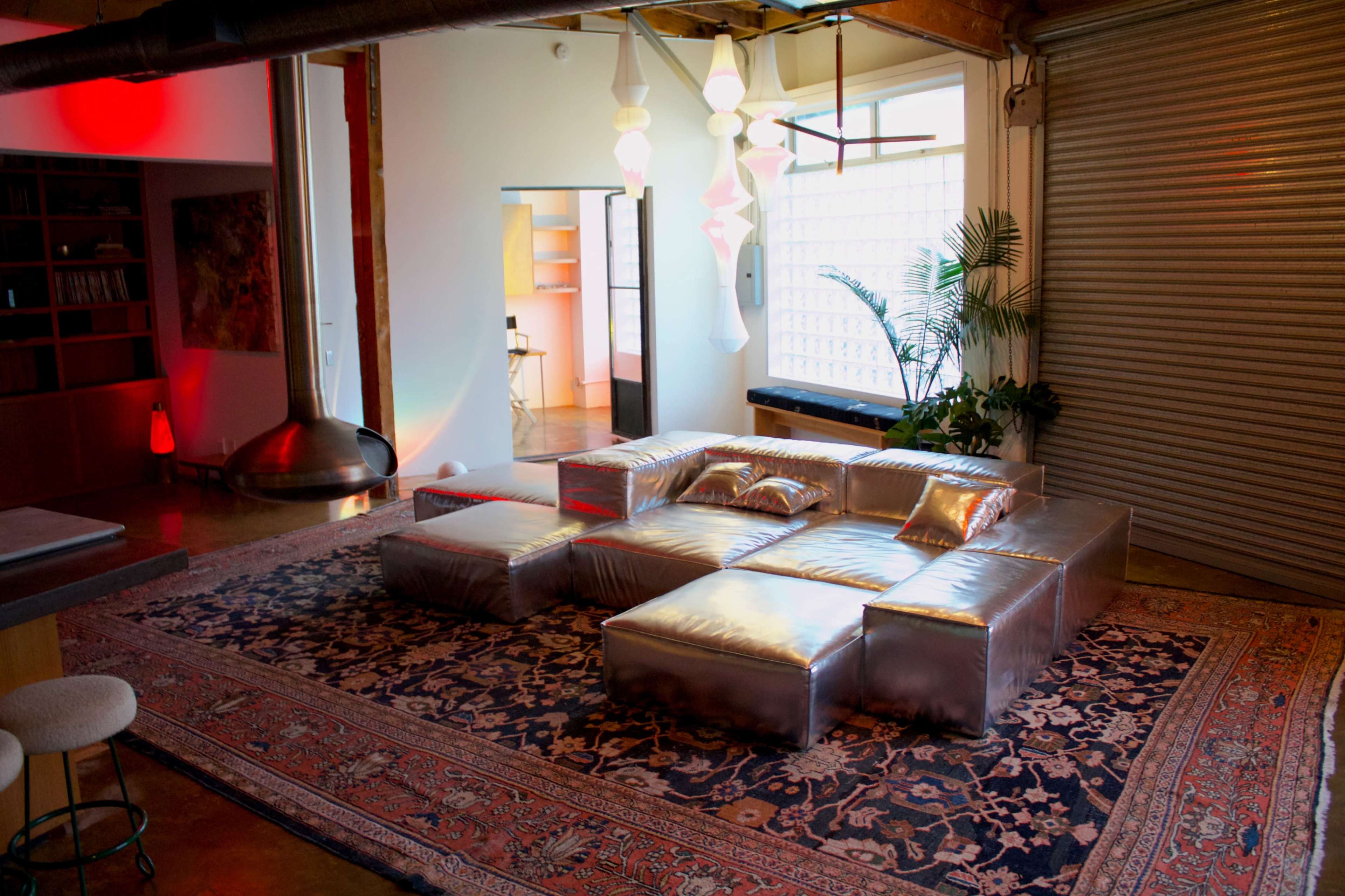 The image shows a modern living space featuring a silver modular seating arrangement on a patterned rug, with plants and stylish lighting fixtures.