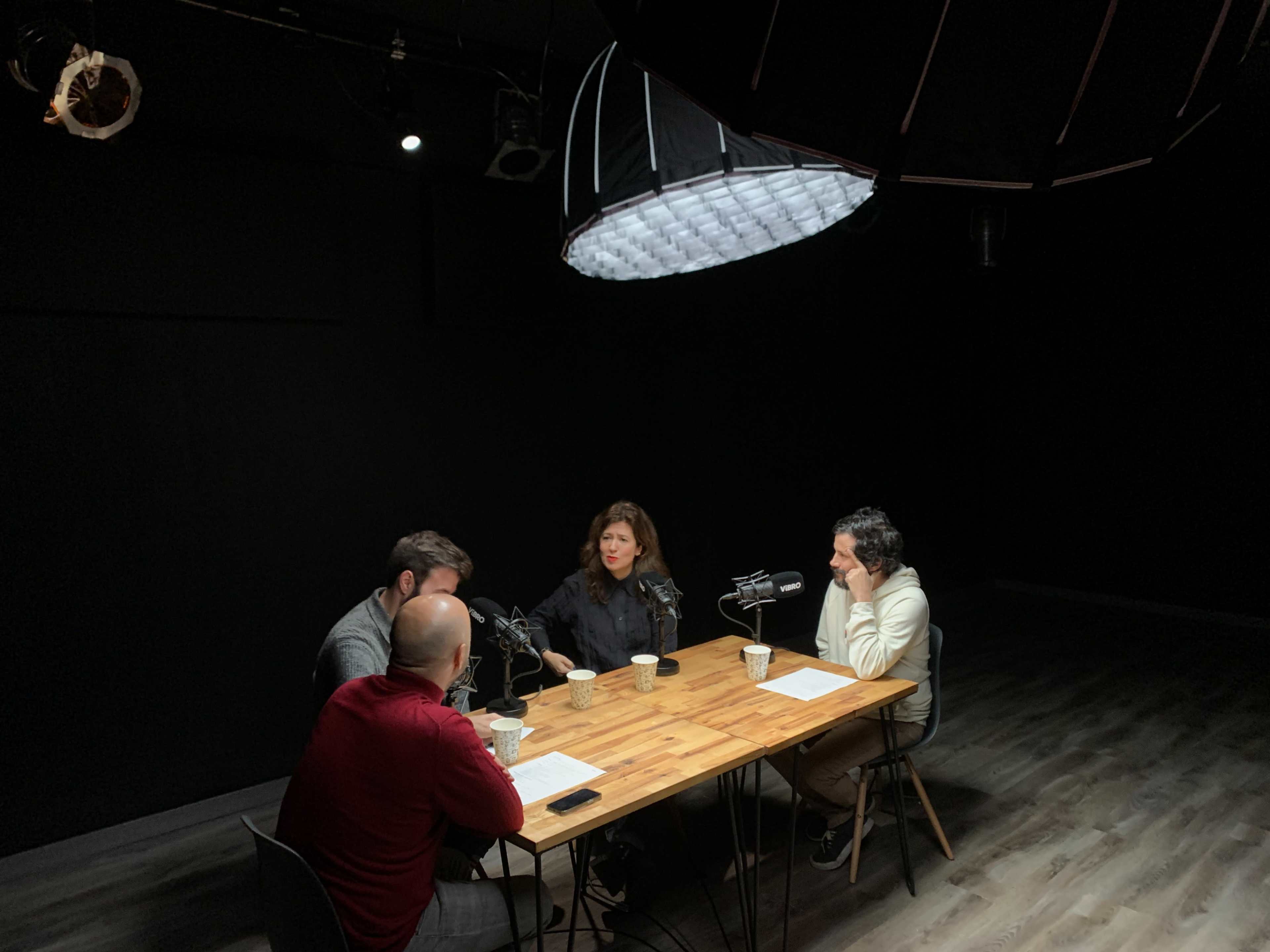 Four people are seated around a wooden table in a dimly lit studio, each with a microphone and coffee cups in front of them.