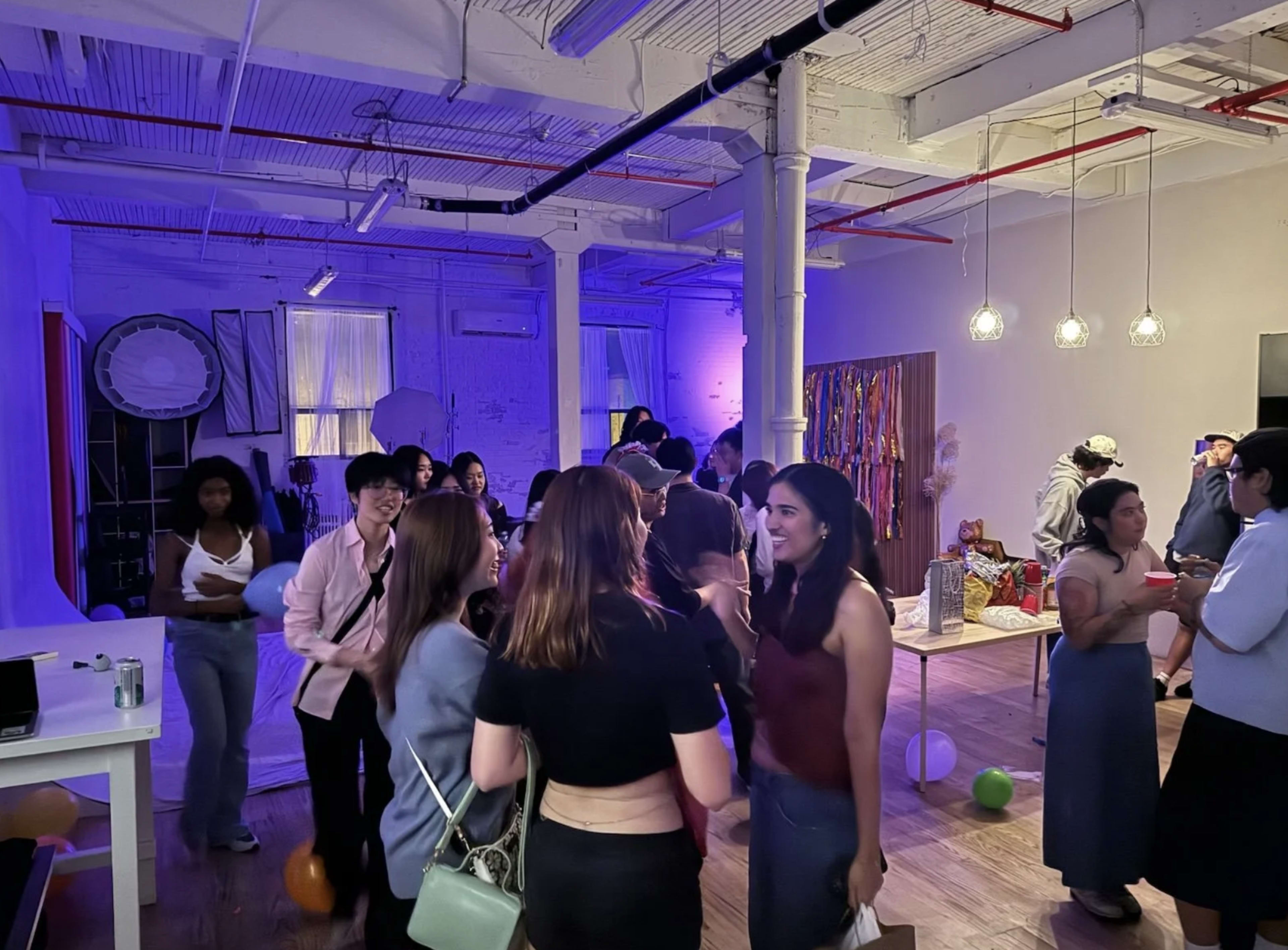 A group of people are socializing and mingling at a brightly lit event in a spacious room decorated with colorful streamers and balloons.