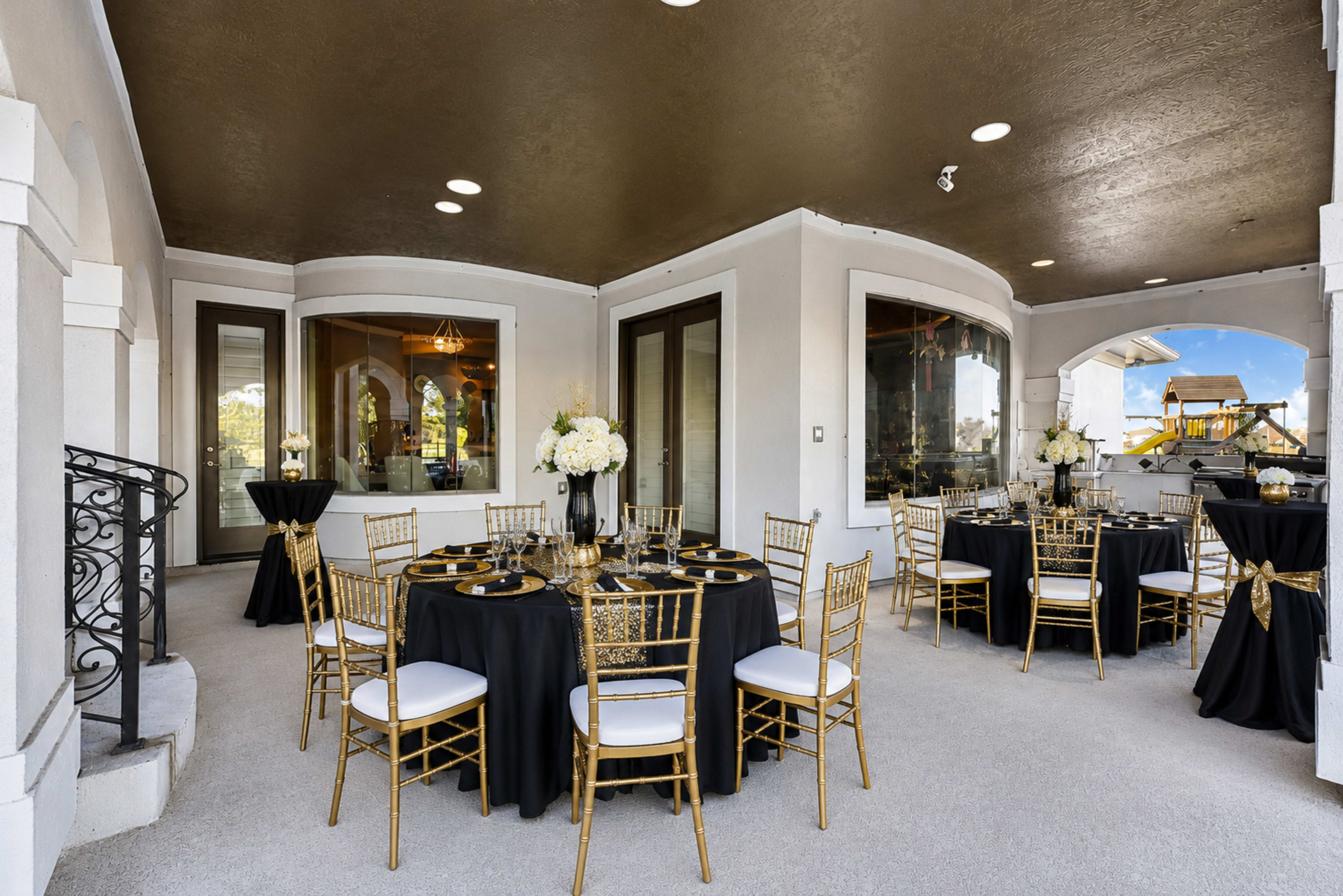 The image shows an elegantly arranged outdoor dining area with round tables covered in black tablecloths and adorned with gold accents and floral centerpieces.
