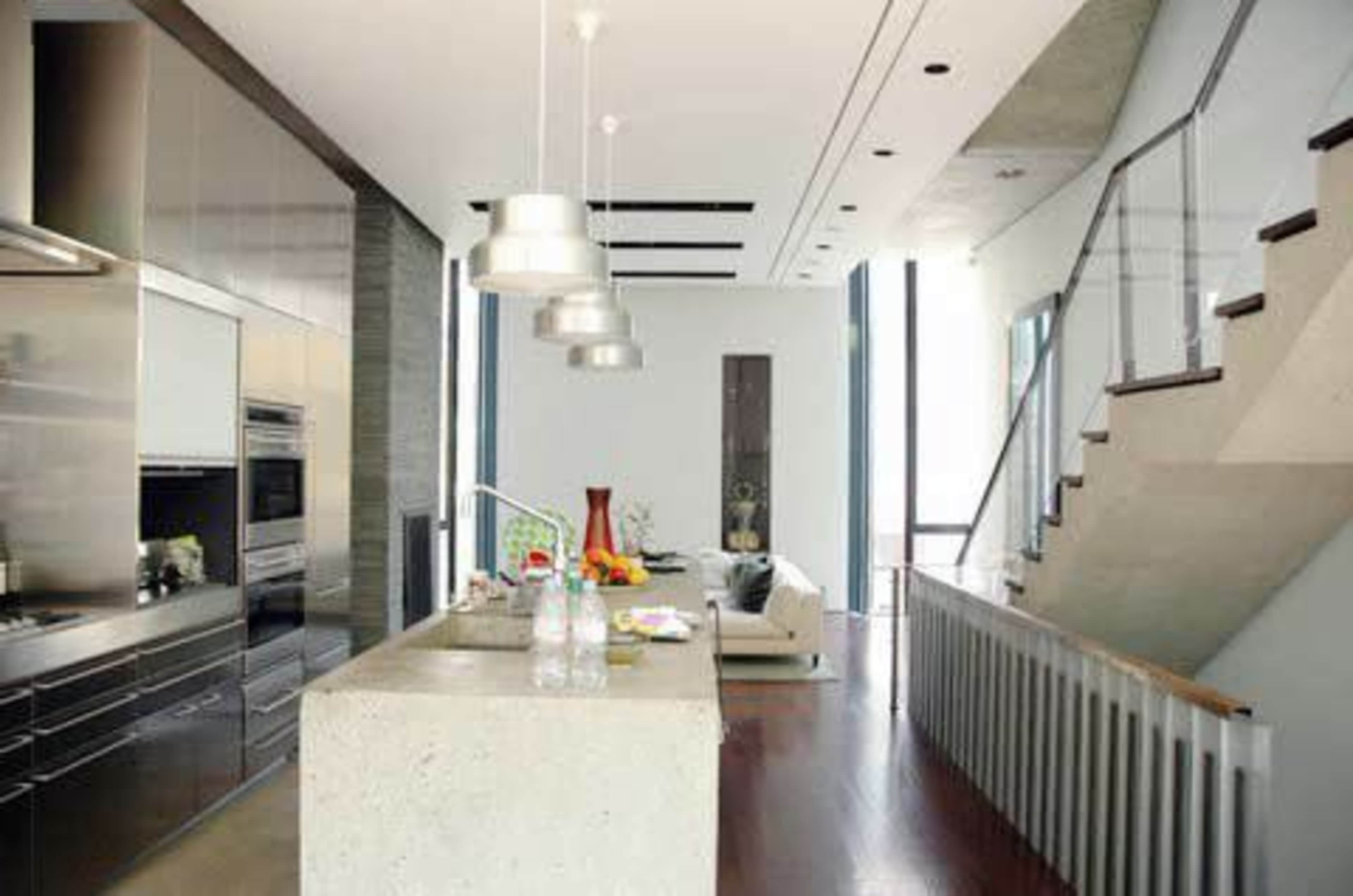 A modern kitchen with a concrete island and stainless steel appliances, adjacent to a living area and a staircase.