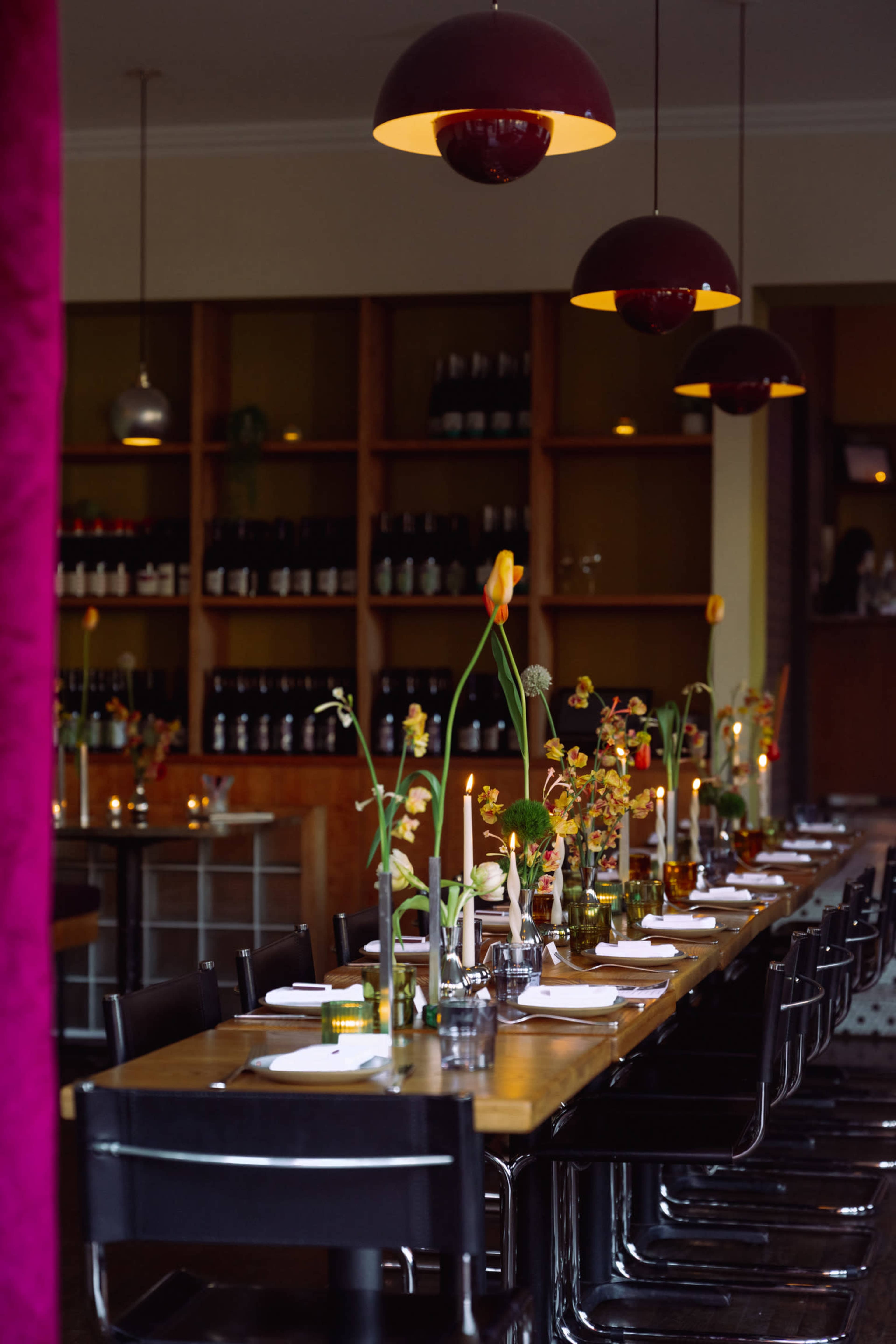 A long wooden dining table is set with flowers and tableware, surrounded by black chairs, beneath pendant lights, in a restaurant with shelves of wine in the background.
