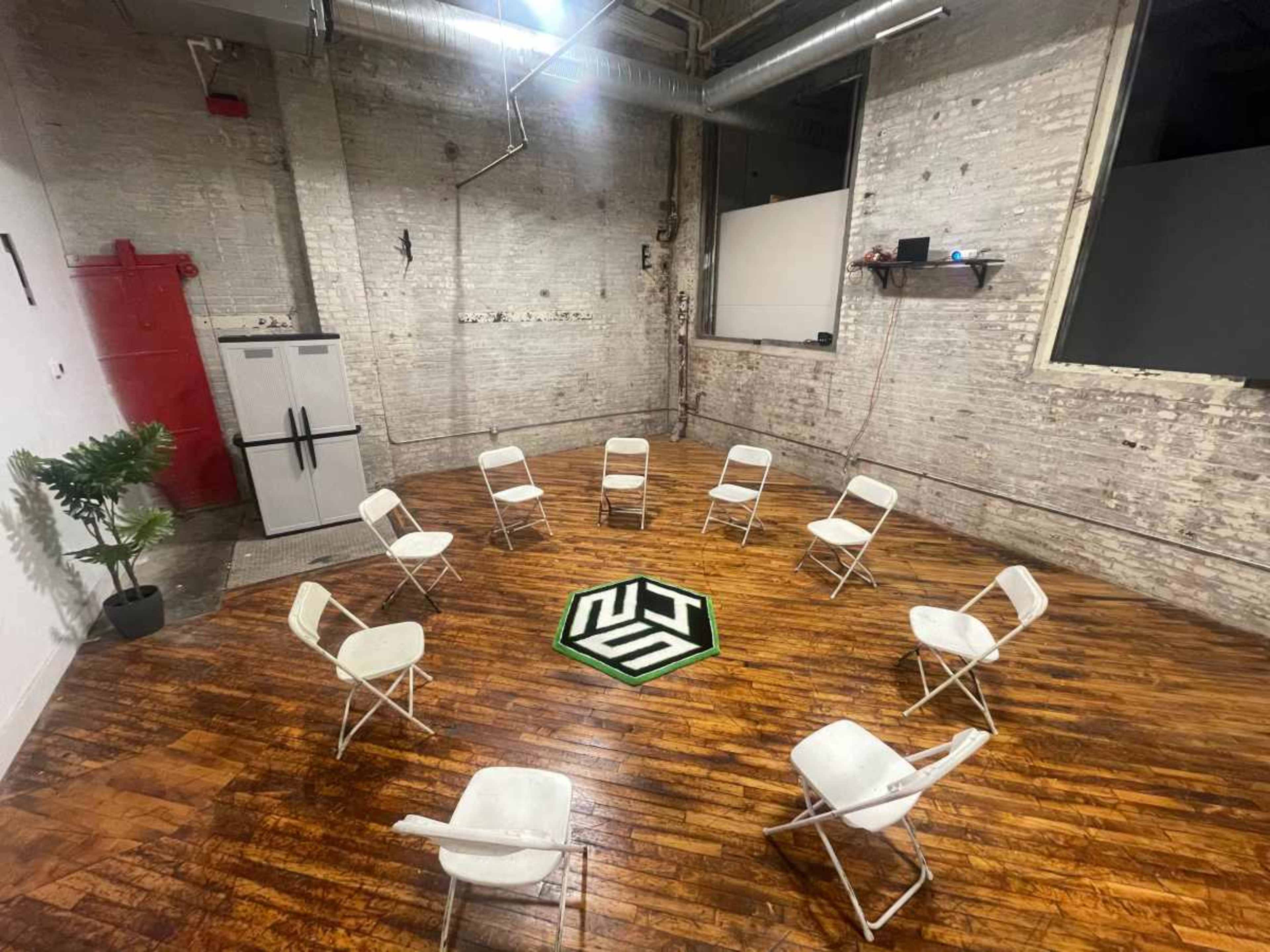 A circular arrangement of white chairs is set on a wooden floor in a sparse, industrial-style room with exposed brick walls and a visible fridge.