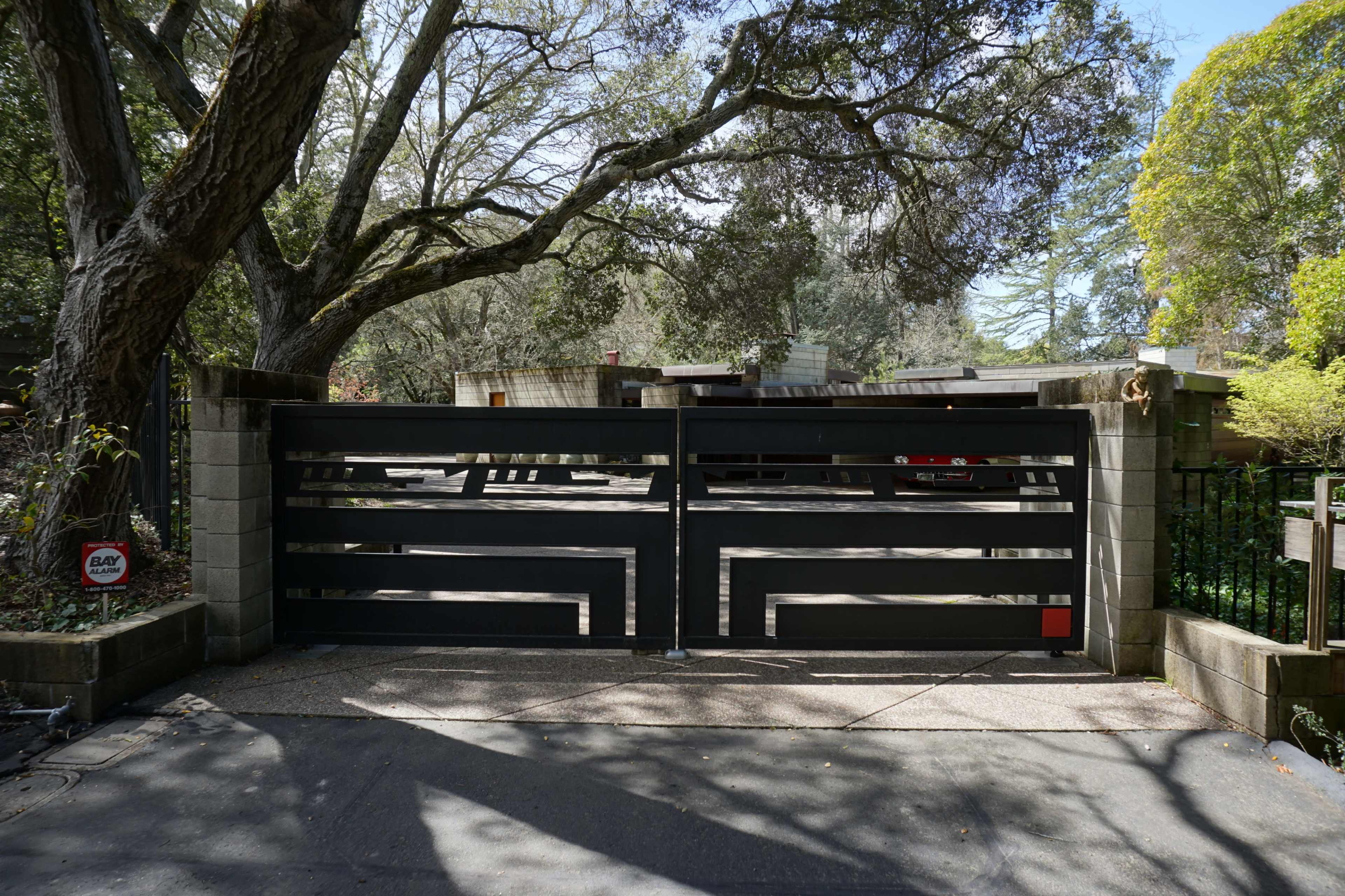 A modern black gate stands closed in front of a property, framed by large trees and a concrete wall.
