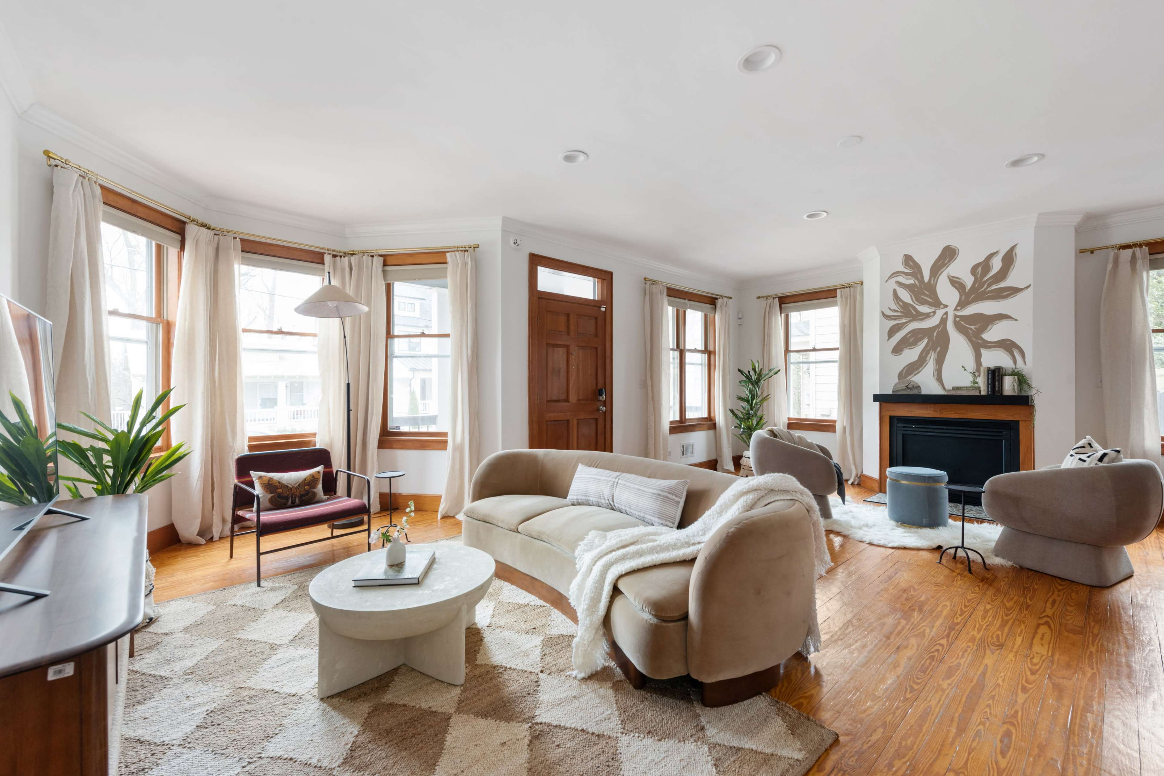A stylish living room features a curved sofa, a circular coffee table, and large windows allowing natural light, with a decorative wall element and a fireplace.