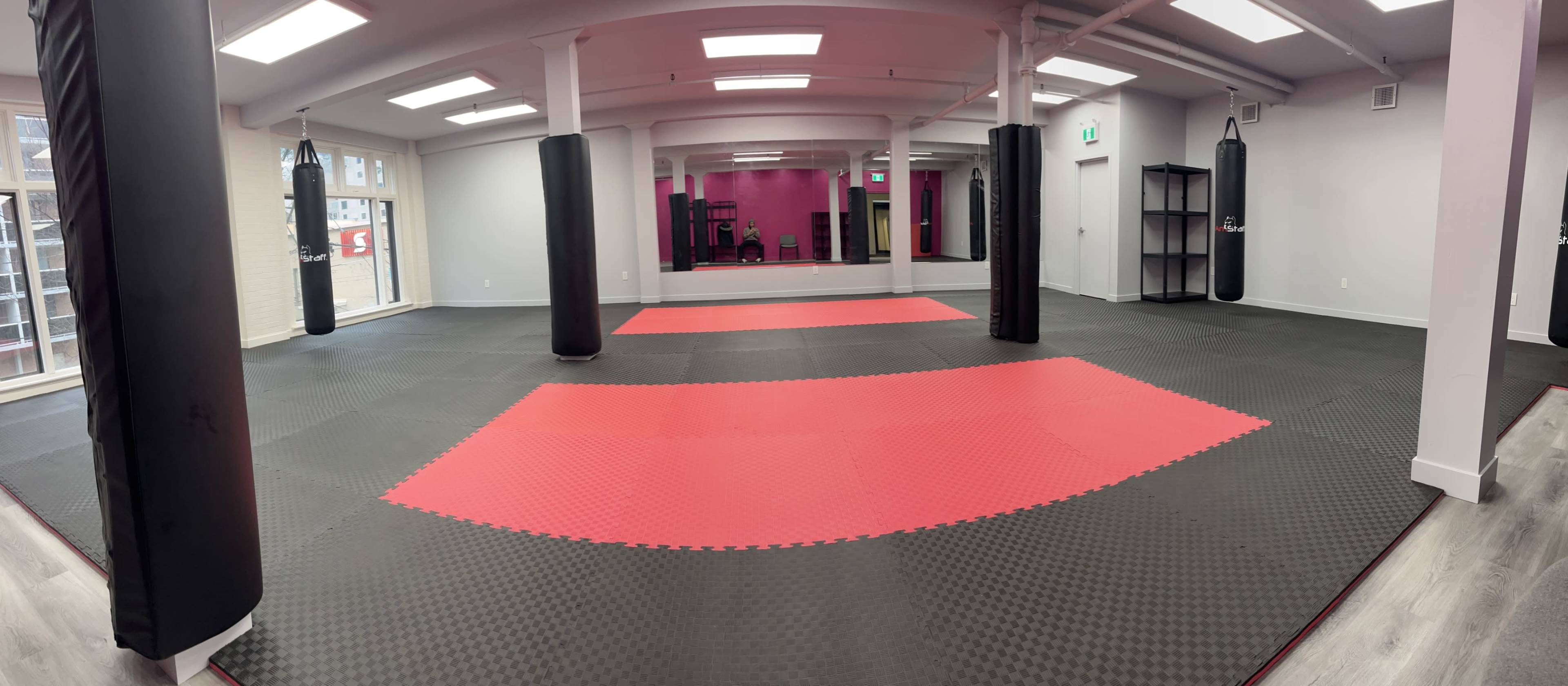 The image shows a spacious, empty martial arts studio with black and red mat flooring and black punching bags positioned around the perimeter.