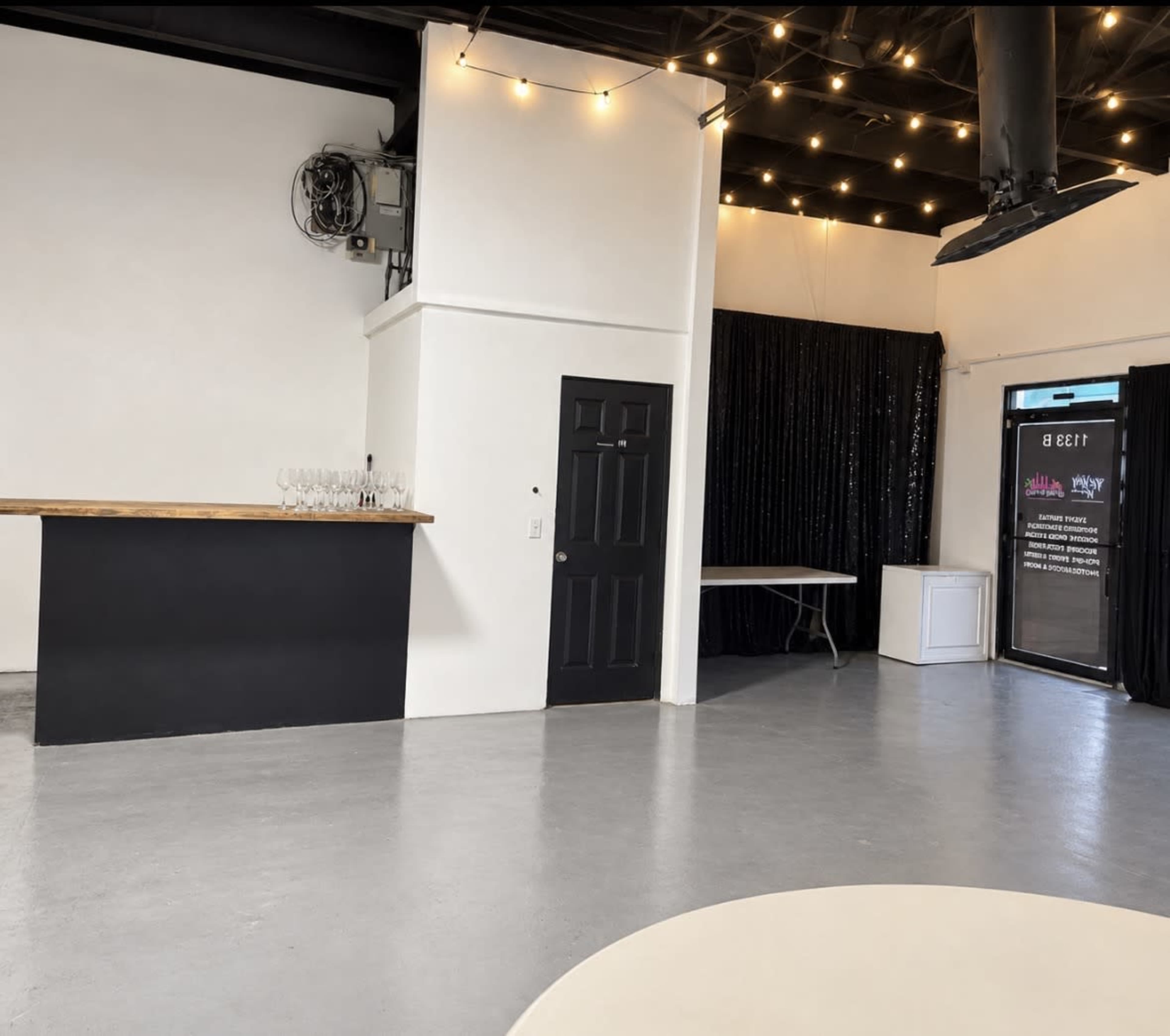 The image shows a spacious indoor venue with a black bar area, a white table, and a large glass door leading outside, surrounded by black drapes and string lights.