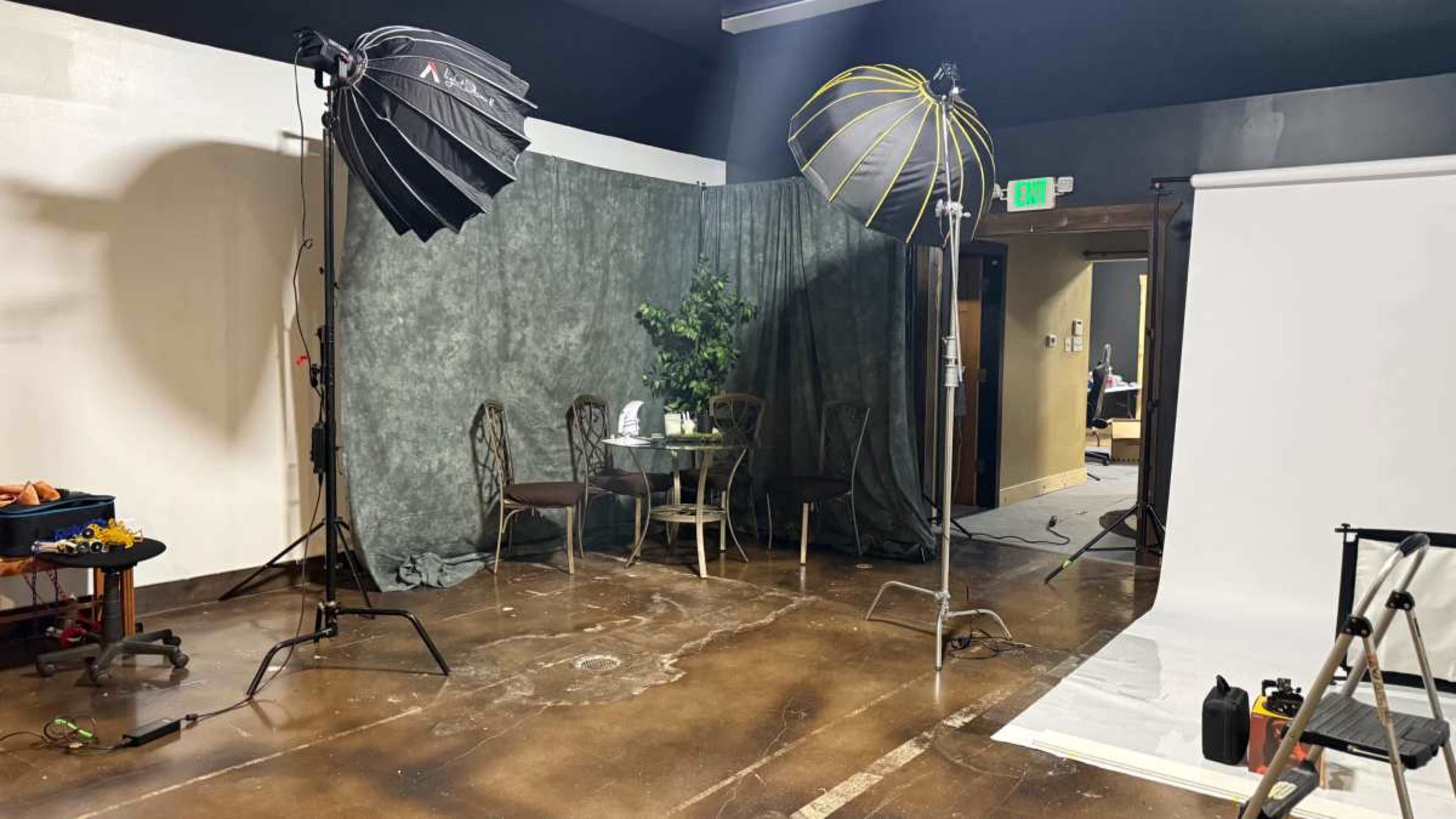 The image shows a photography studio setup featuring two large softboxes, a backdrop of green fabric, a small table with chairs, and a white photography backdrop in the corner.