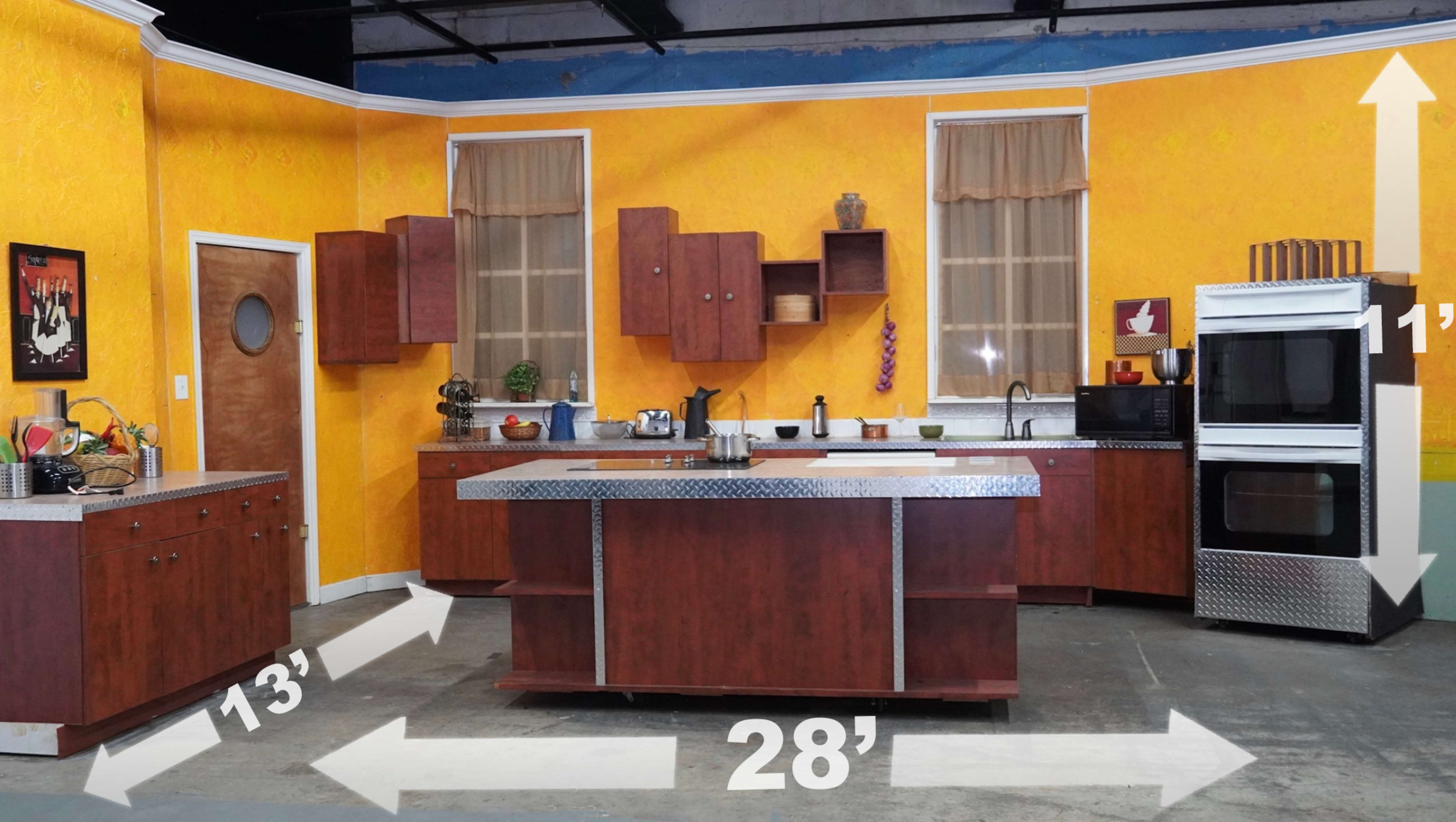 The image features a kitchen set with wooden cabinets, a central island, and various appliances, all situated against a bright yellow wall.