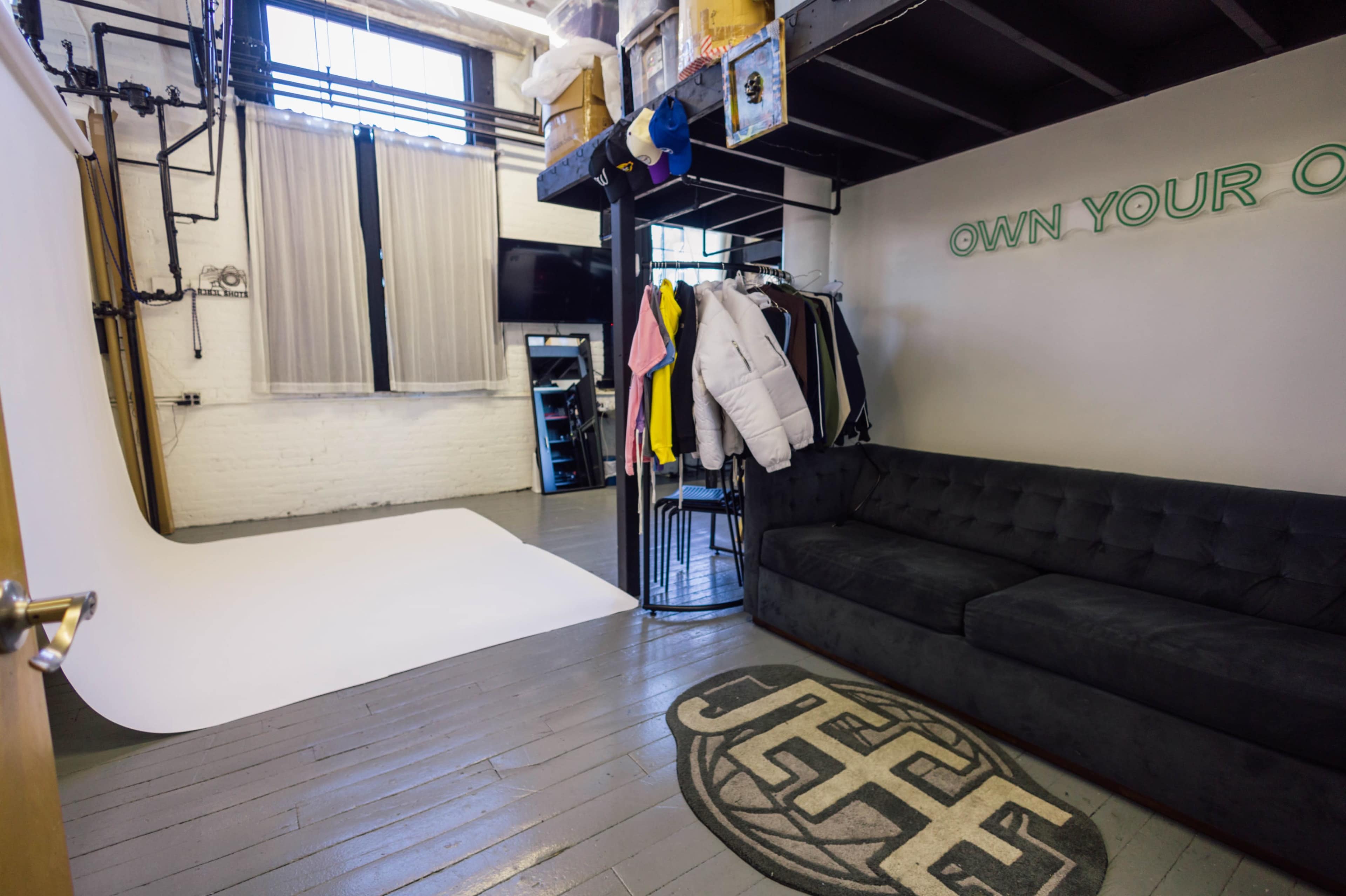 The image depicts a photography studio featuring a white backdrop, a black sectional couch, clothing racks, and a wall displaying the phrase "OWN YOUR OWN."
