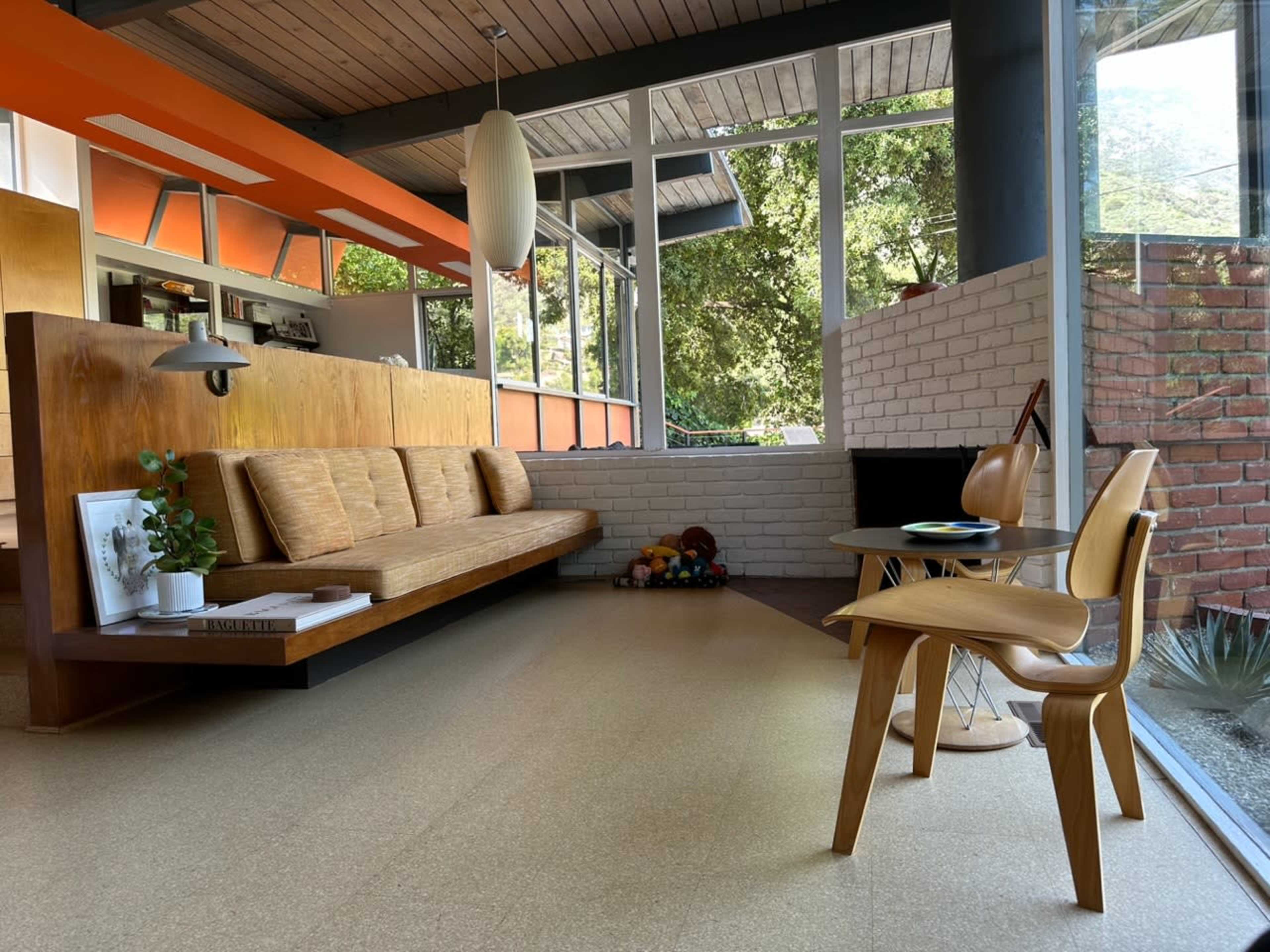 A modern interior with a wooden sofa, a chair, and large windows allowing natural light to enter, surrounded by greenery.