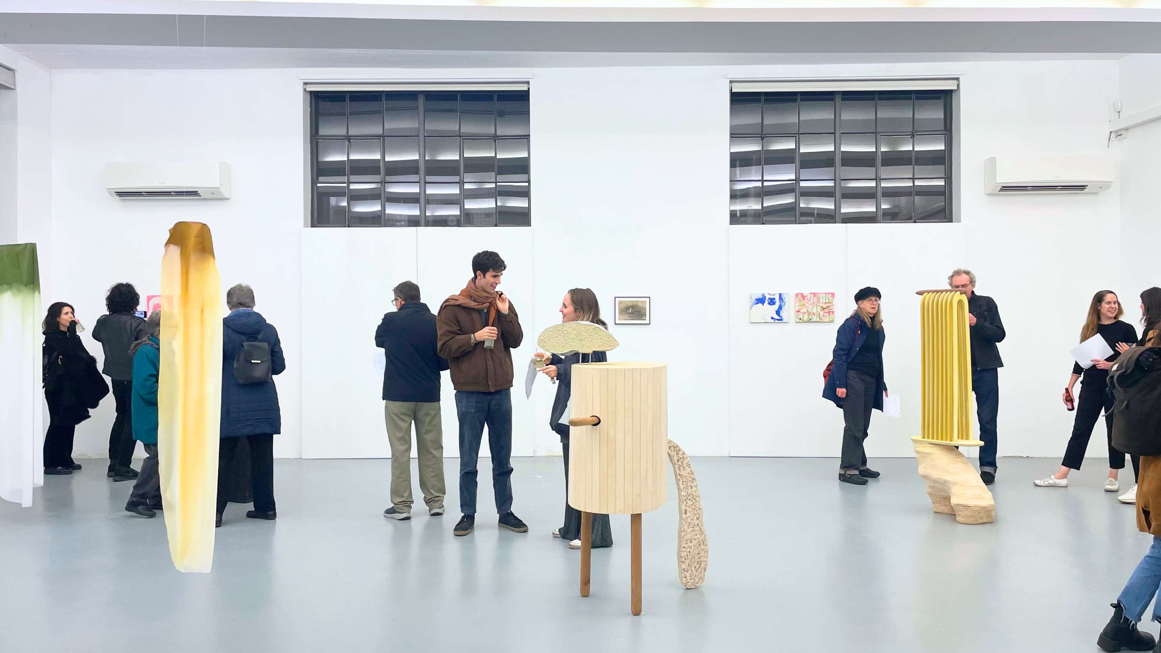 A contemporary art gallery features attendees interacting with various artistic displays, including sculptures and framed artworks on the walls.