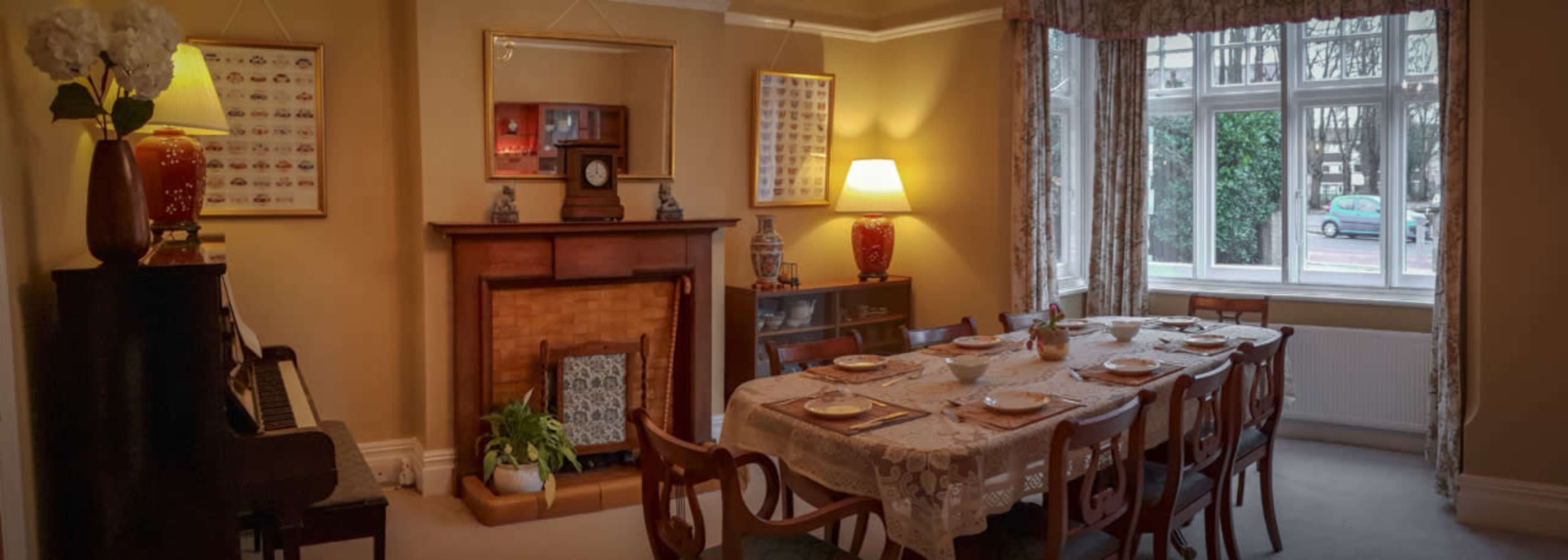 A dining room with a large table set for a meal, surrounded by wooden chairs, a piano in the corner, and a window offering a view outside.