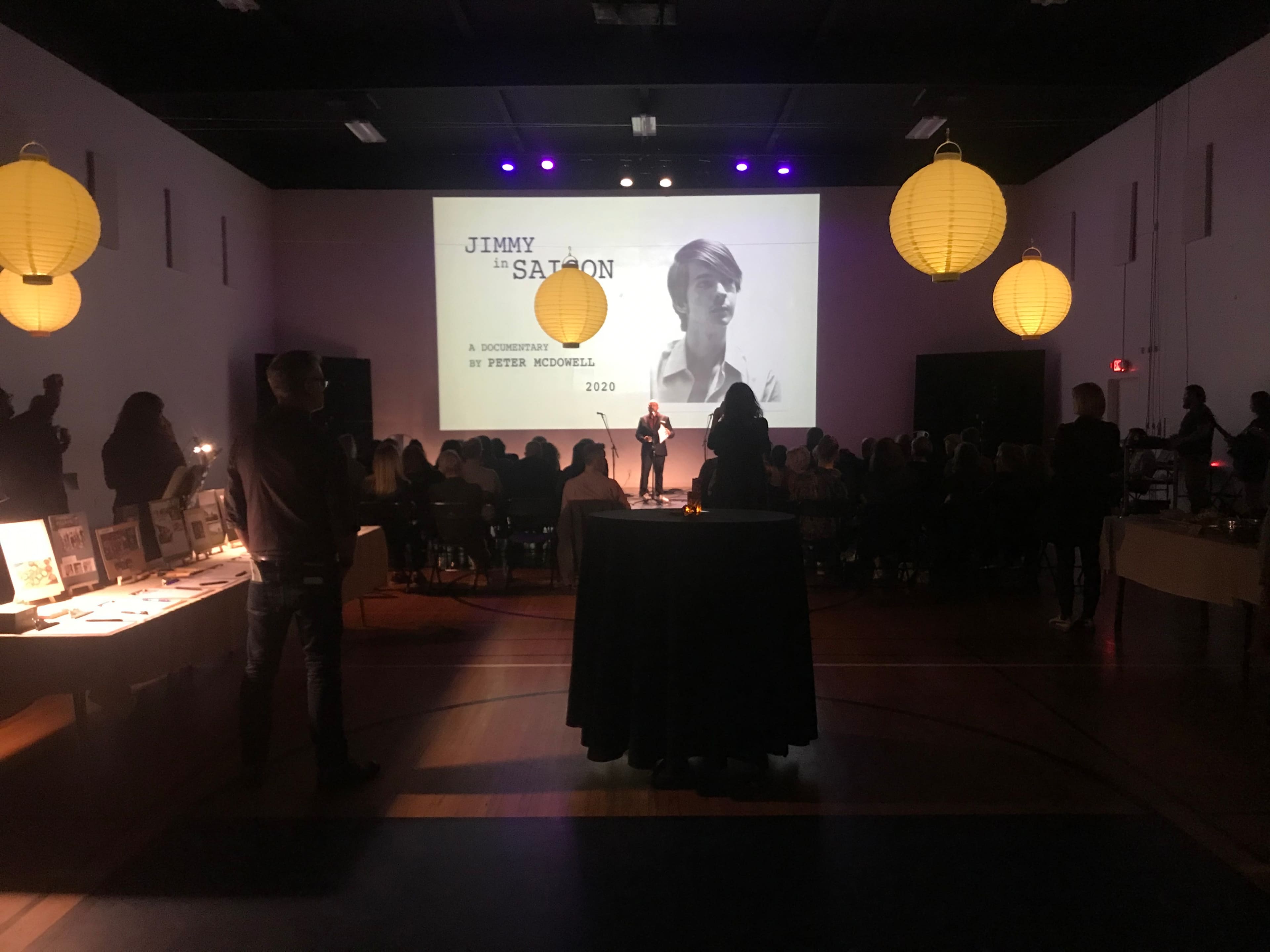 A projector screen displays "JIMMY 'SATION," while a speaker stands on stage in front of an audience amidst glowing lanterns.