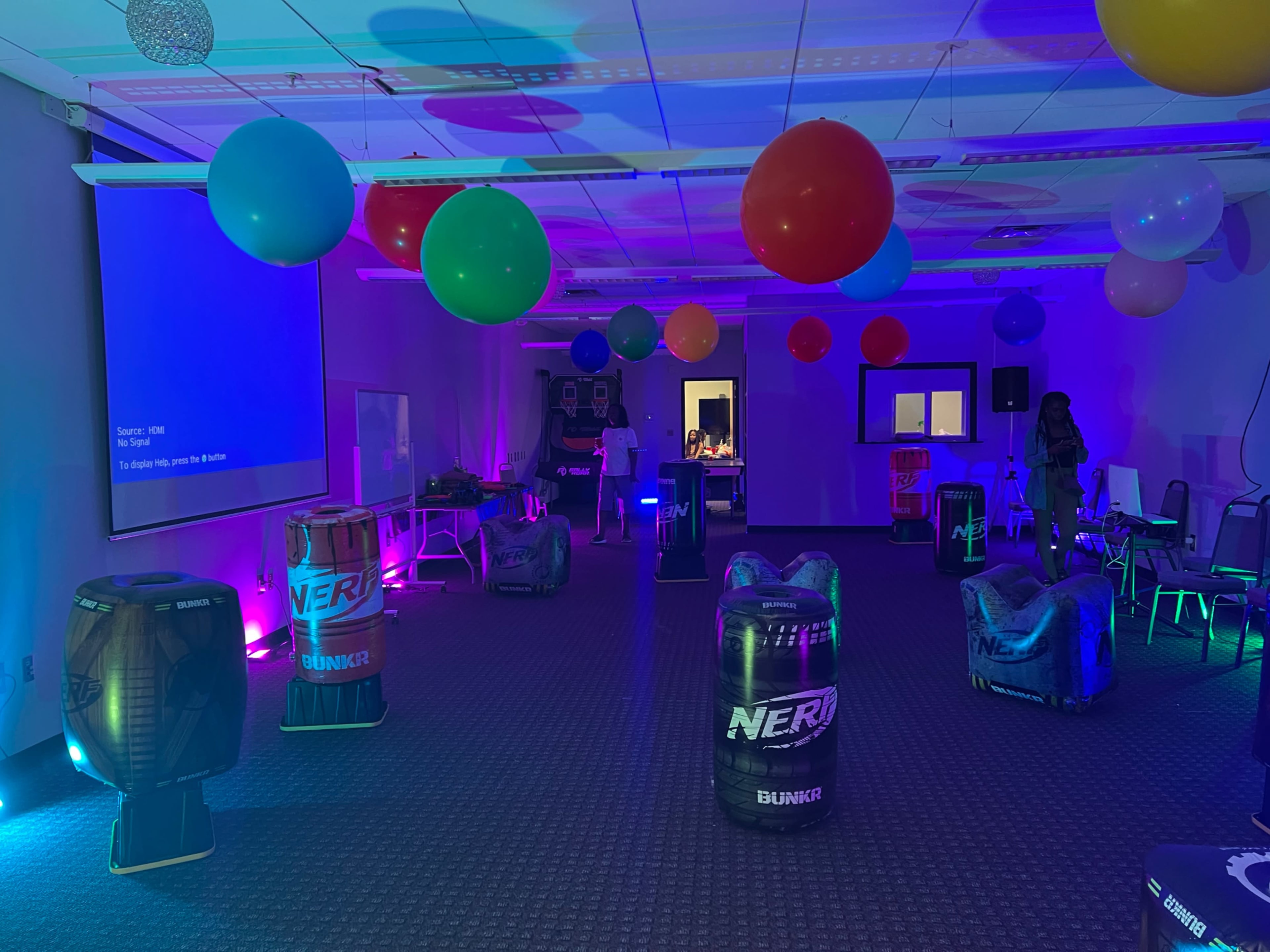 The room is set up for an event, featuring colorful balloons and a series of Nerf-themed obstacles arranged around a carpeted floor.