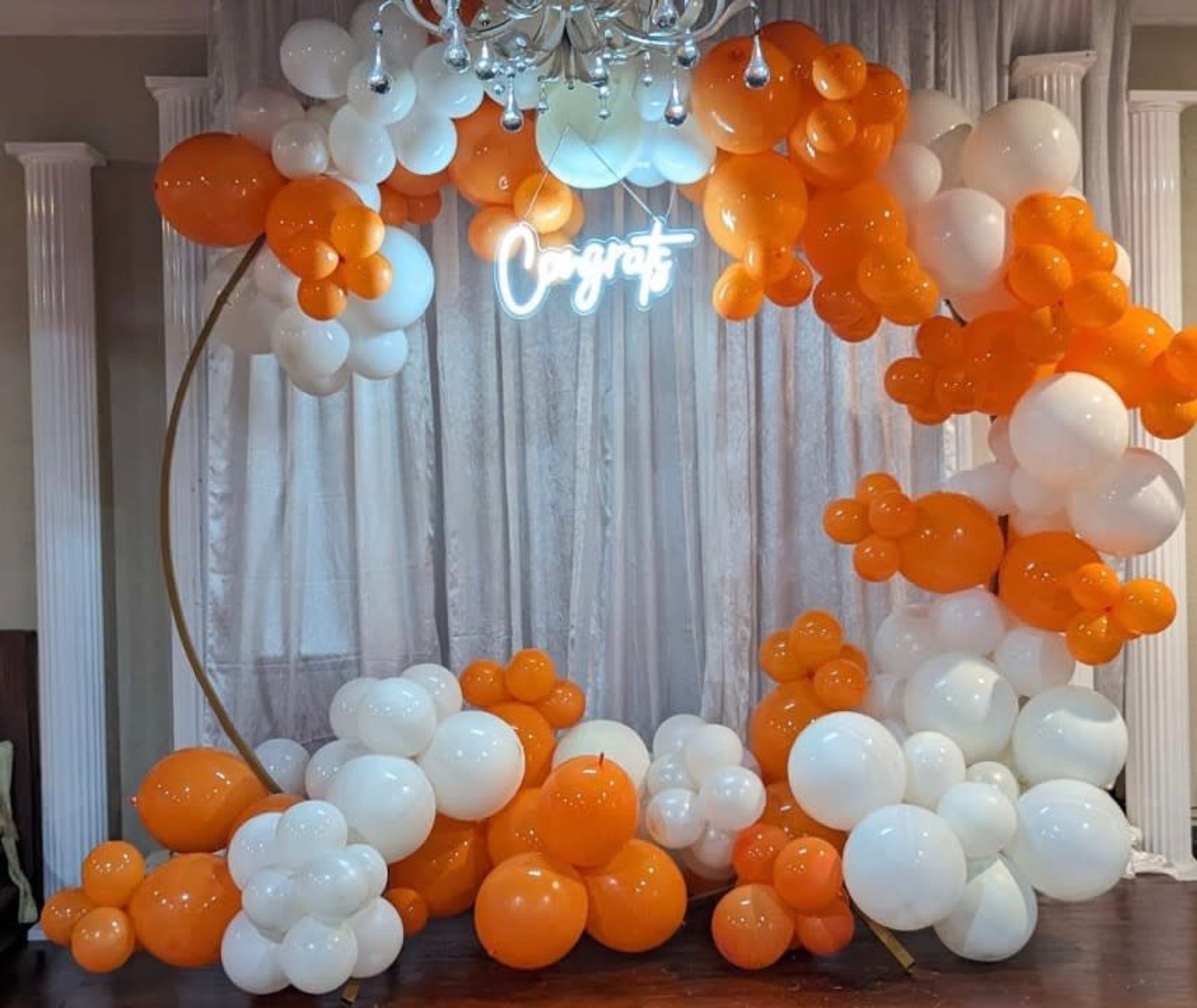 A circular balloon arch decorated with orange and white balloons, with a neon sign that says "Congrats" at the center.