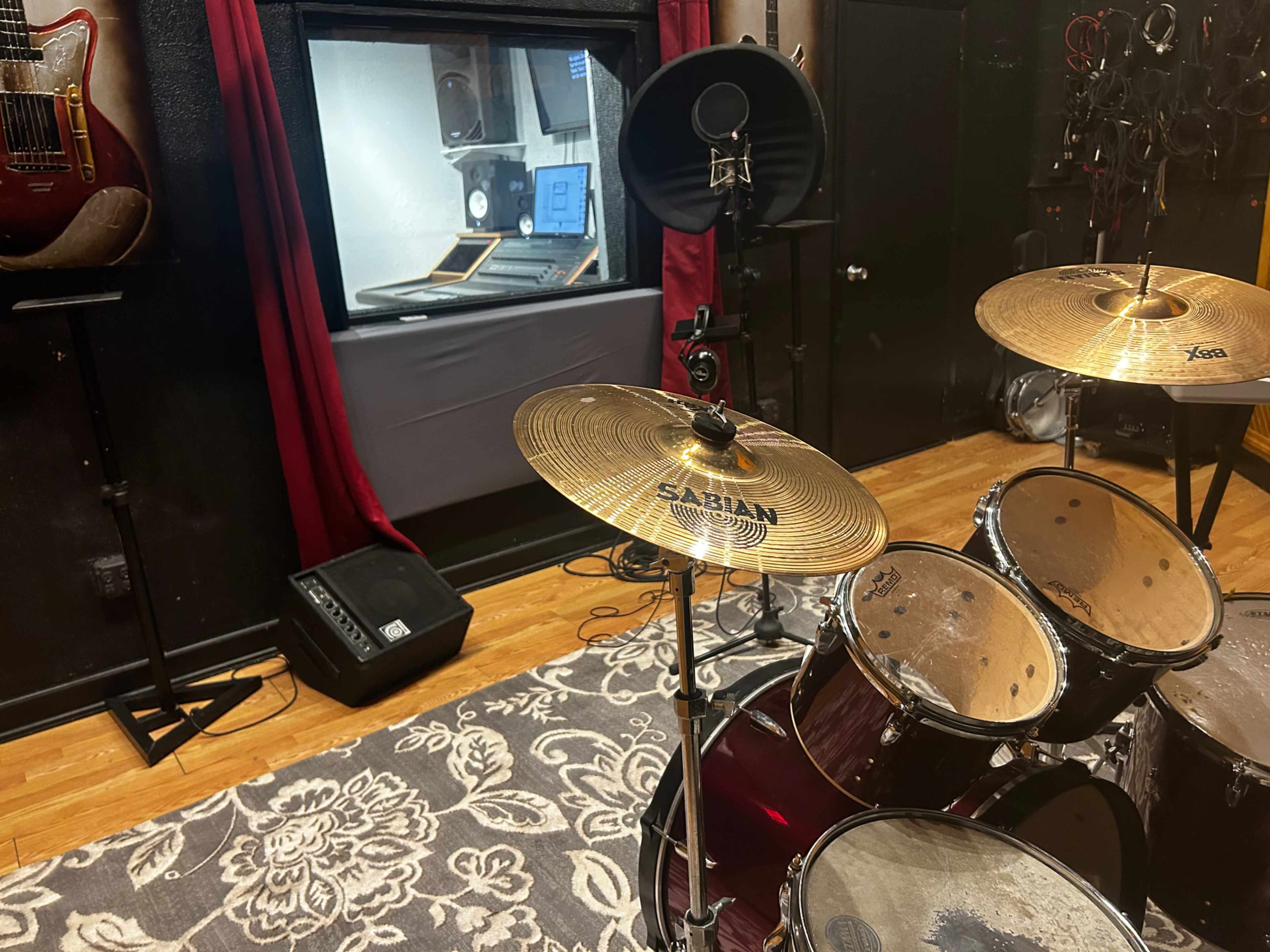 A drum set is positioned in a room that features a soundproof window and recording equipment visible in the background.