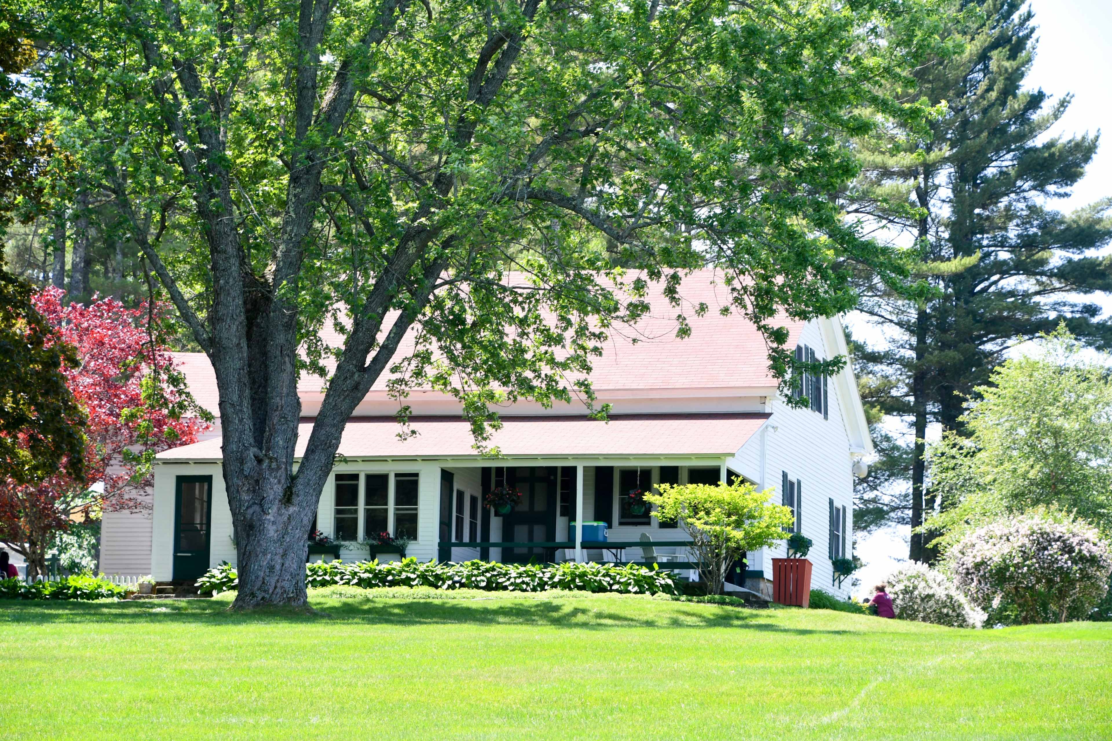 A white two-story house with a porch is situated on a grassy lawn surrounded by trees and flowering shrubs.