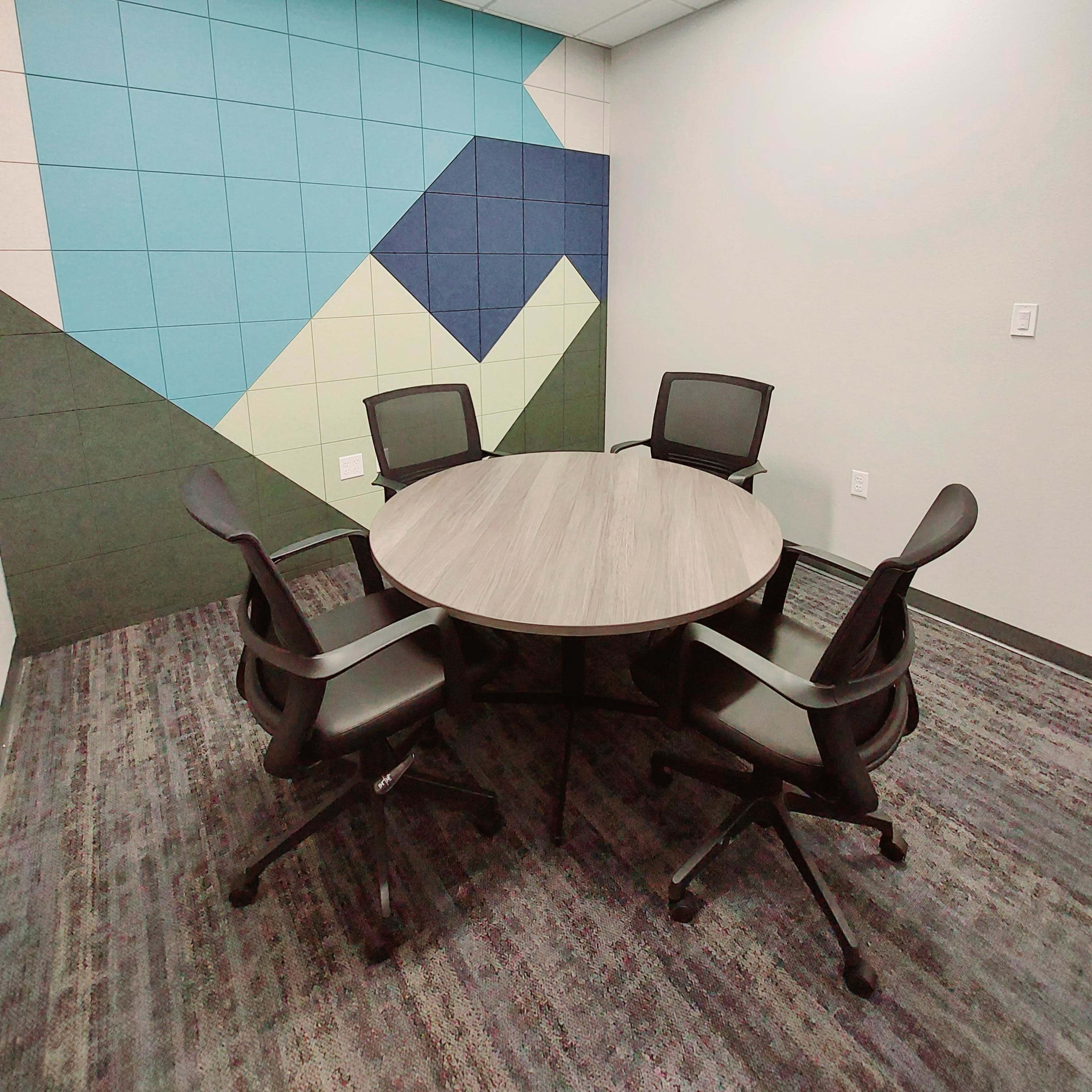 A round table with four black chairs is positioned in a small meeting room featuring a colorful geometric wall.