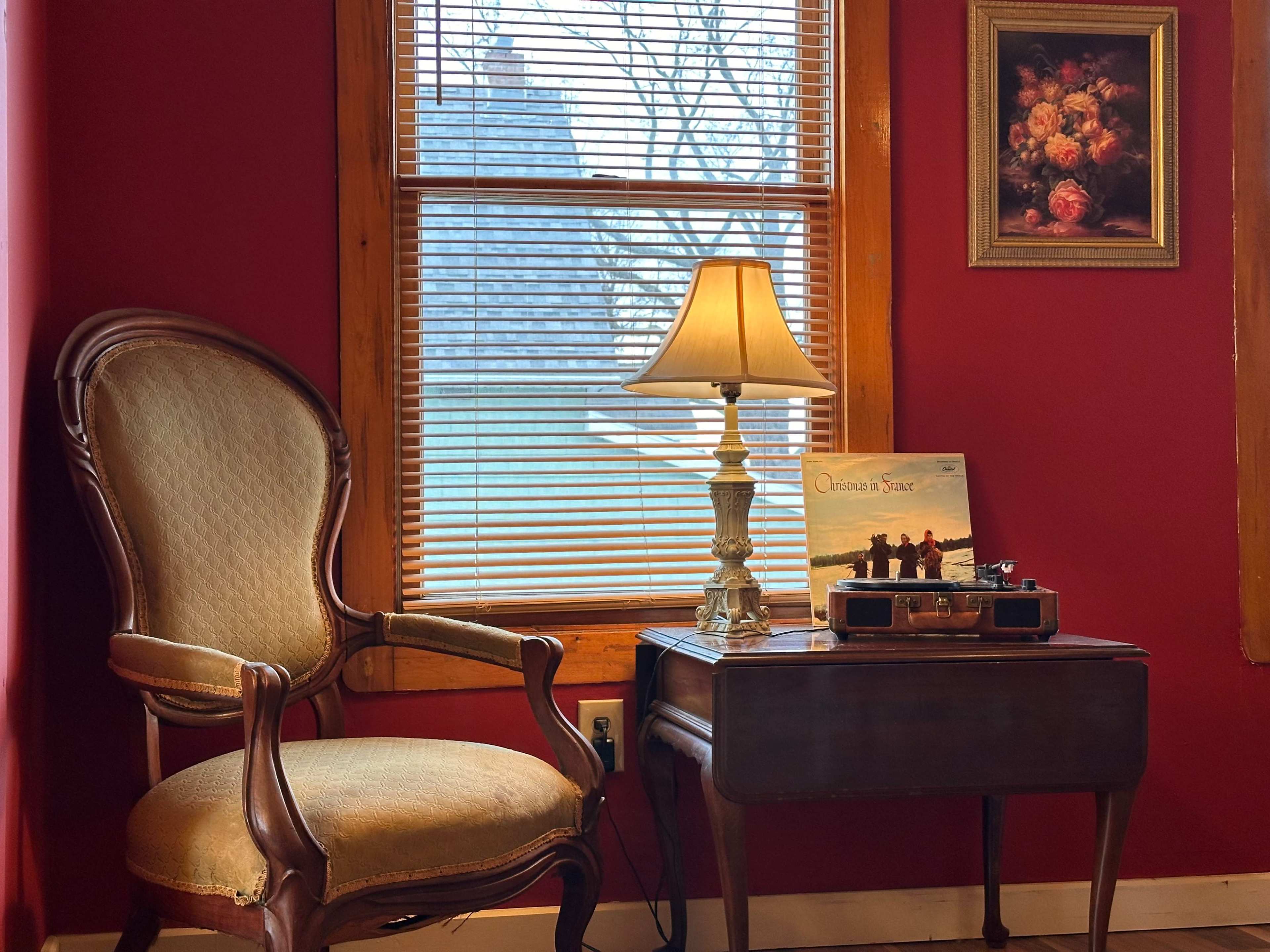A vintage armchair sits beside a small table with a lamp, a record player, and a framed painting on a red wall near a window.