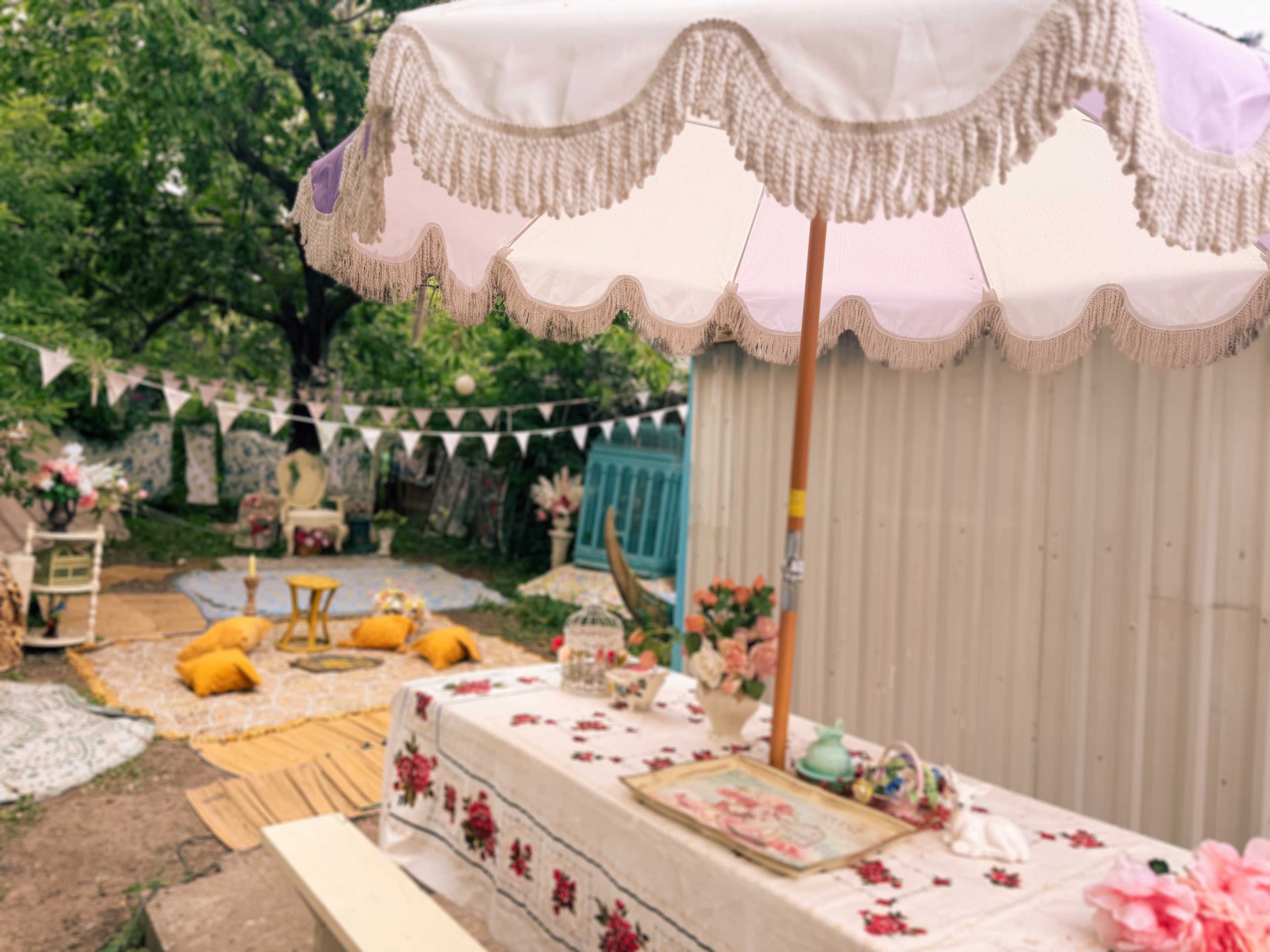 A large, fringed umbrella shades a decorated table set with flowers and tea items, surrounded by colorful cushions and festive bunting in an outdoor garden setting.