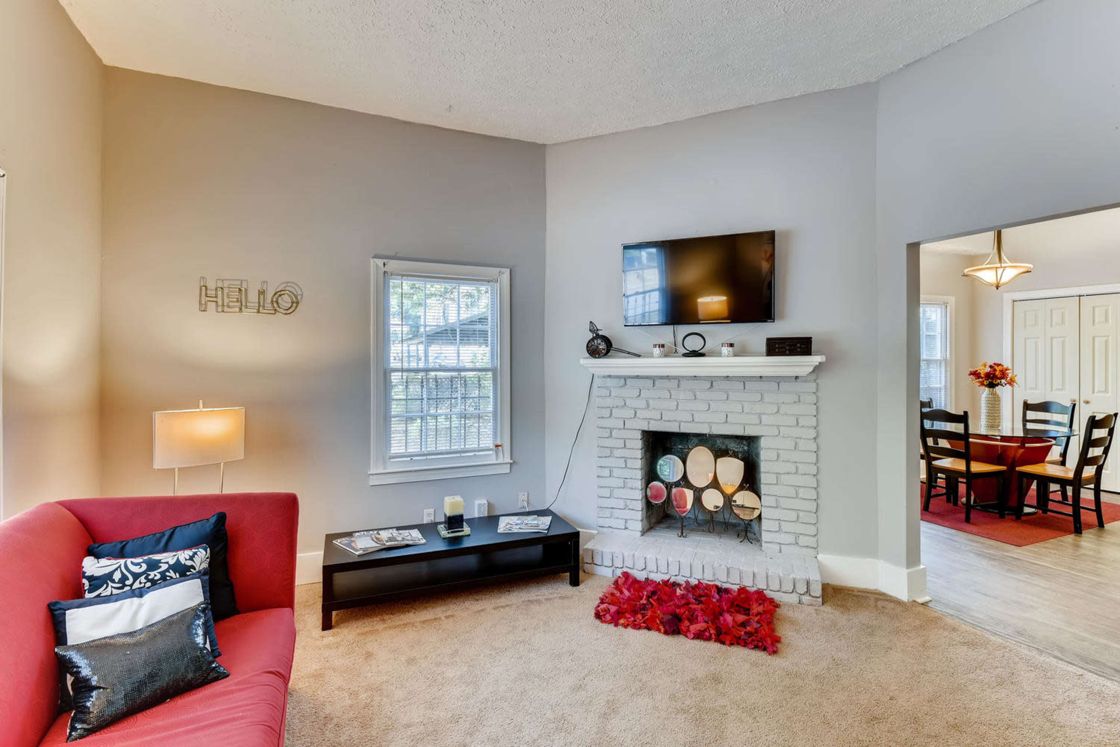 A cozy living room features a red couch, a television mounted above a white brick fireplace, and a dark coffee table, with a dining area visible in the background.