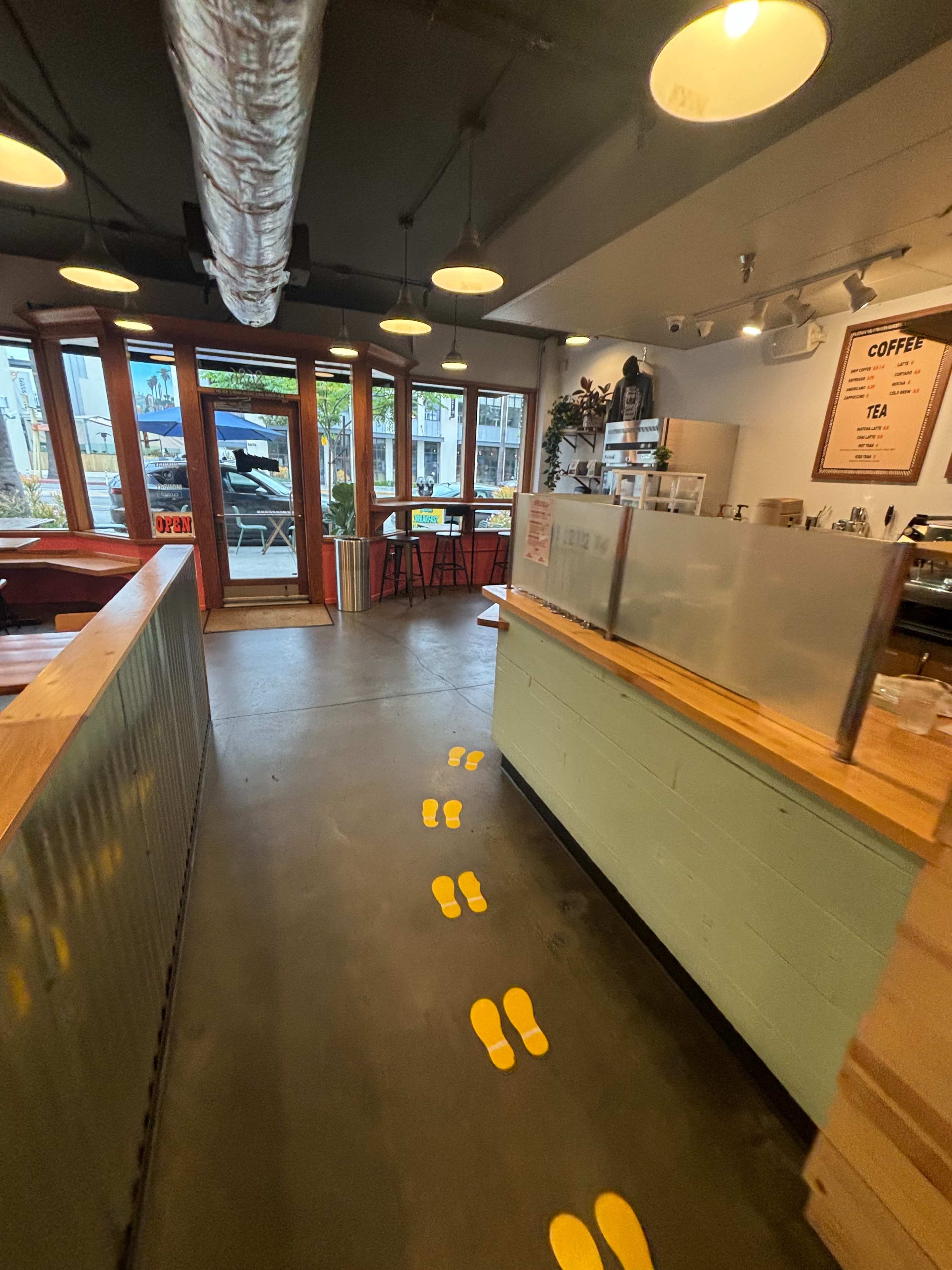 The image shows a café interior with yellow footprints leading from the entrance to the counter, surrounded by wooden furniture and large windows.