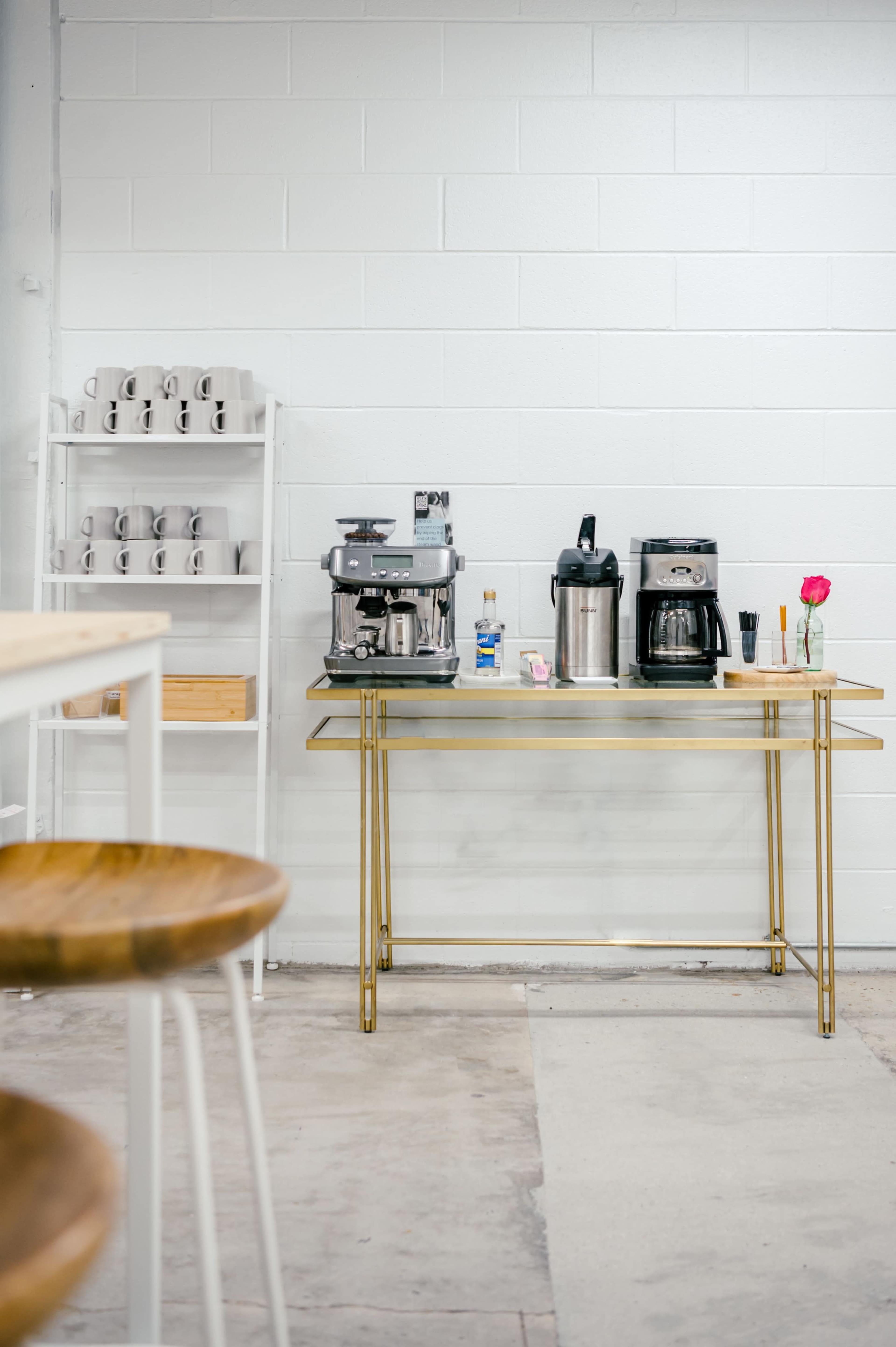 A coffee station featuring an espresso machine, a coffee maker, and a vase with a flower, set against a white wall with shelves of mugs.