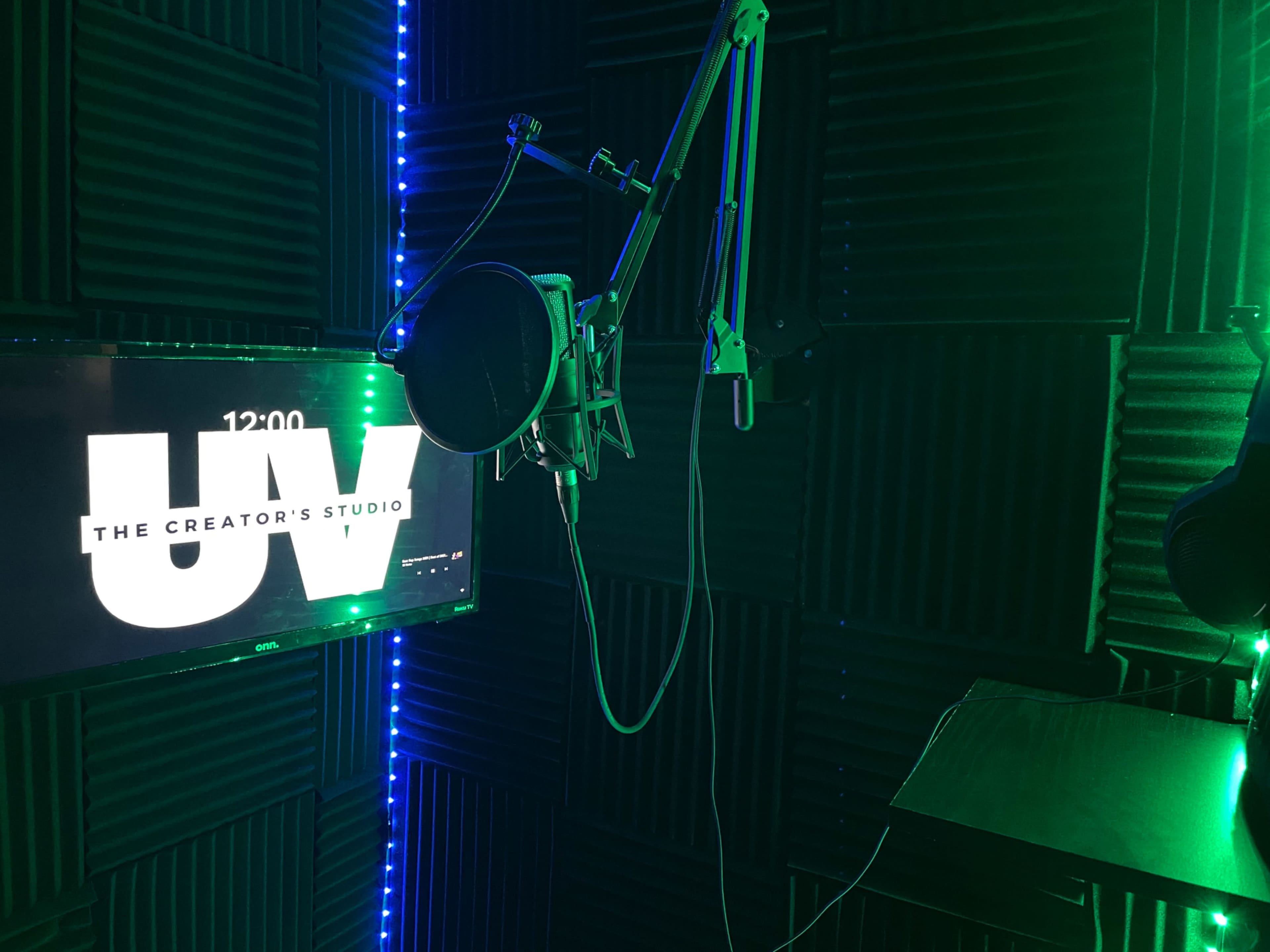 A sound studio with green lighting, featuring a microphone mounted on an adjustable arm and a large display screen labeled "UV" and "THE CREATOR'S STUDIO."