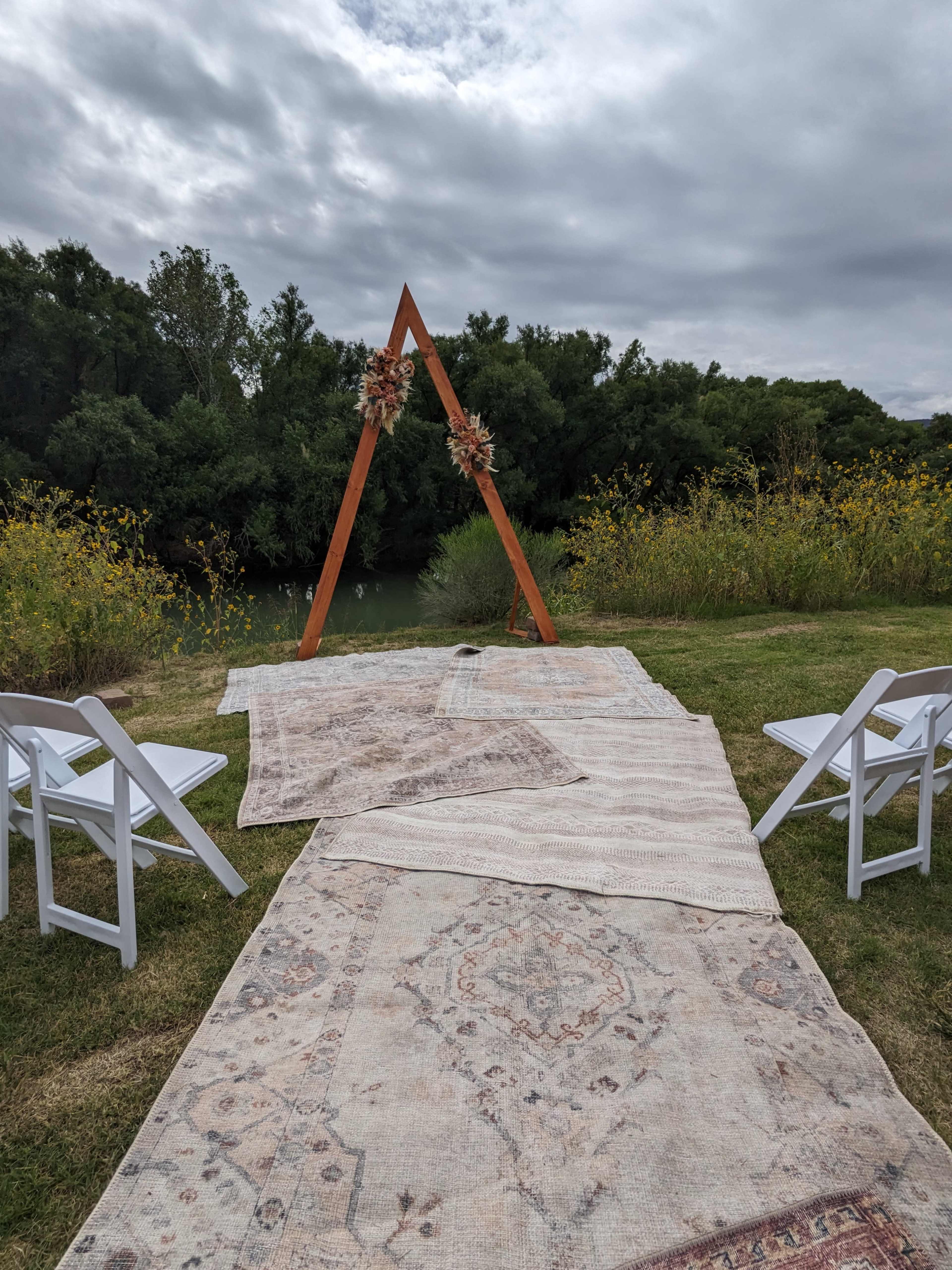 The image shows a rustic outdoor wedding setup featuring an A-frame structure adorned with flowers, with an aisle made of patterned rugs leading to it, flanked by white chairs.