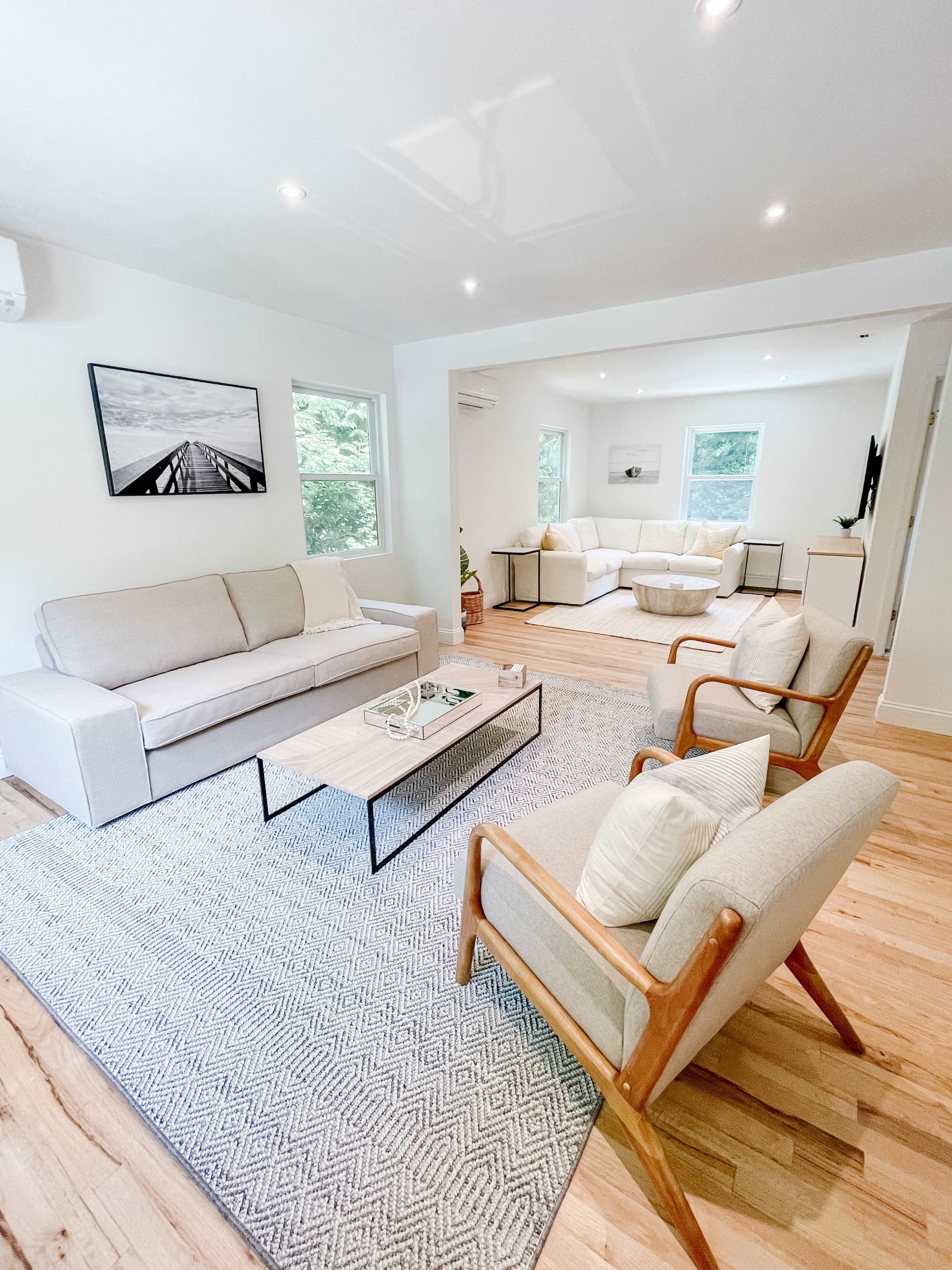 A bright living room with two sofas, a coffee table, and two armchairs arranged on a wooden floor with a patterned rug.