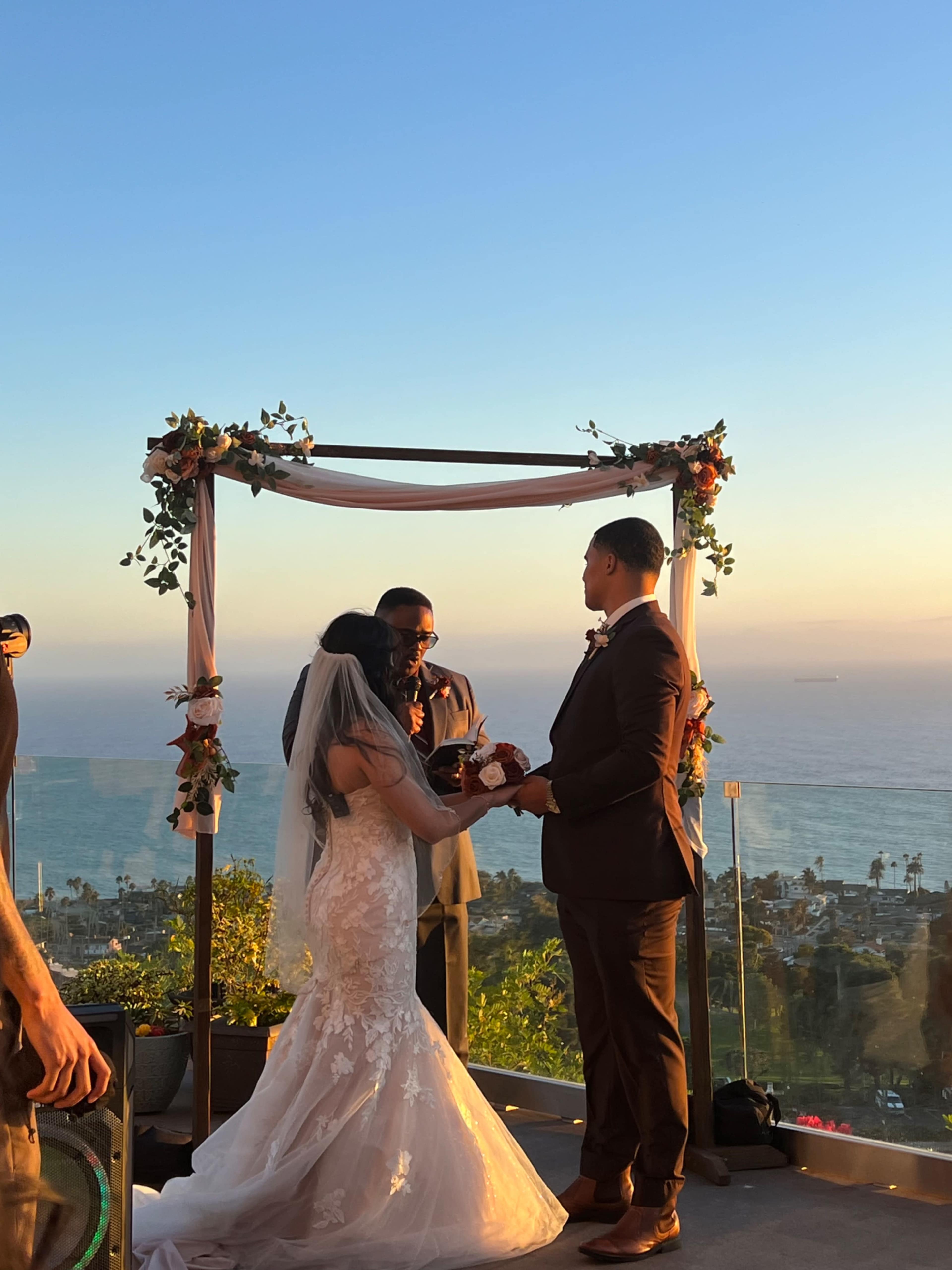 A couple exchanges vows during their wedding ceremony under an arch overlooking the ocean at sunset.