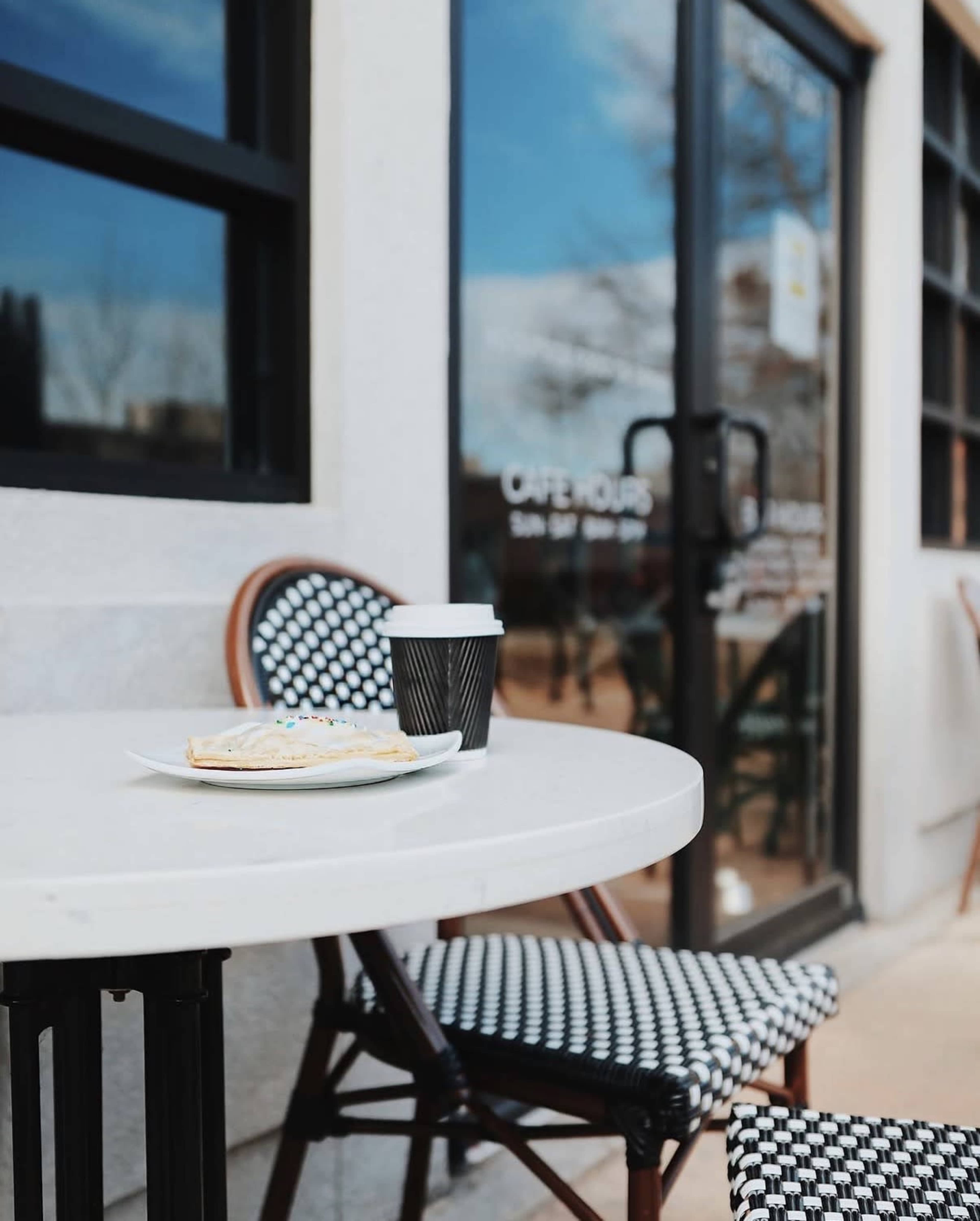 A small table with a plate of pastry and a takeout coffee cup is set outside a café.