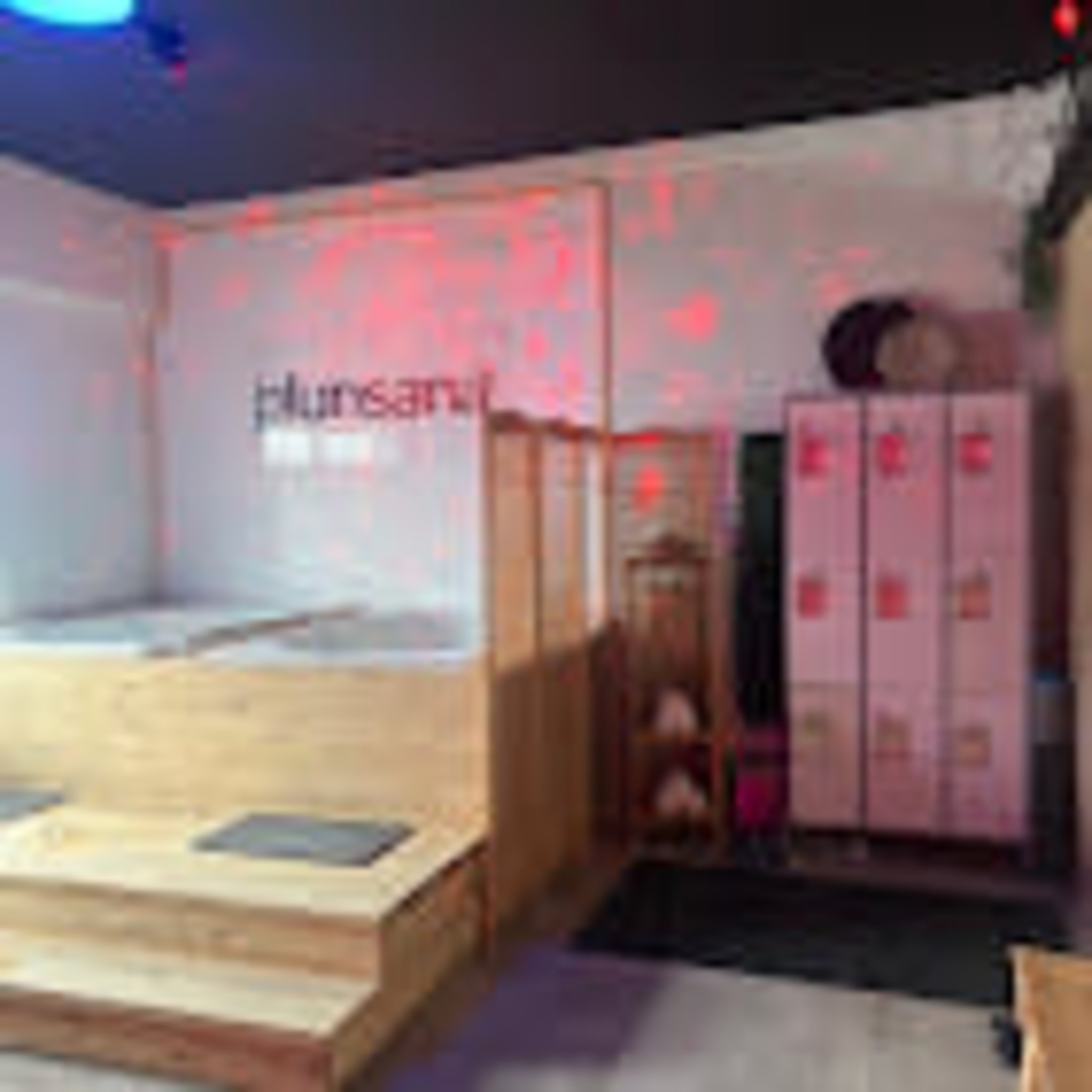 The image shows a spa area featuring wooden seating, lockers, and a wall with the words "plunsari."