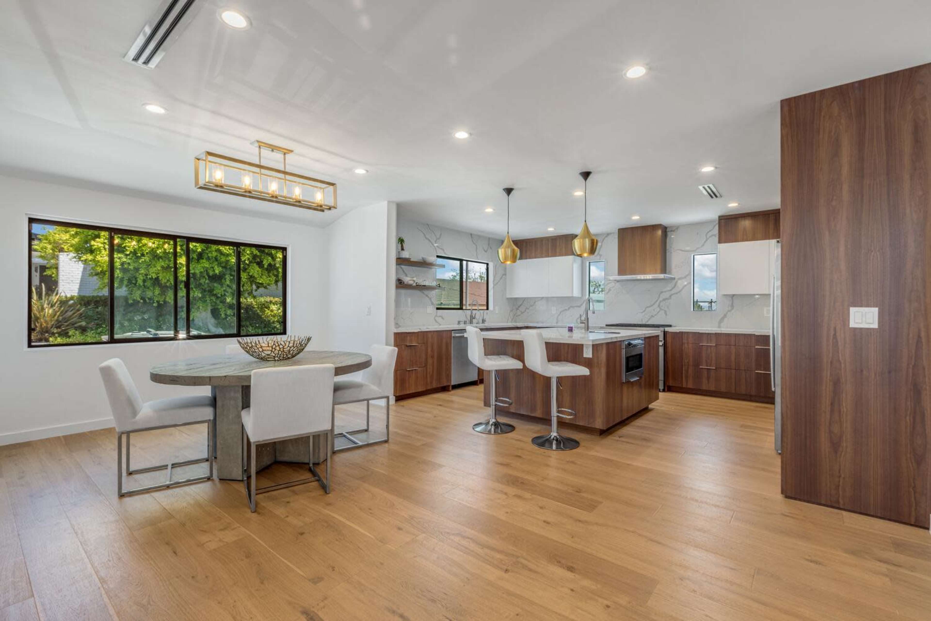 A modern kitchen and dining area featuring a round table, white chairs, and wooden cabinets with large windows allowing natural light.