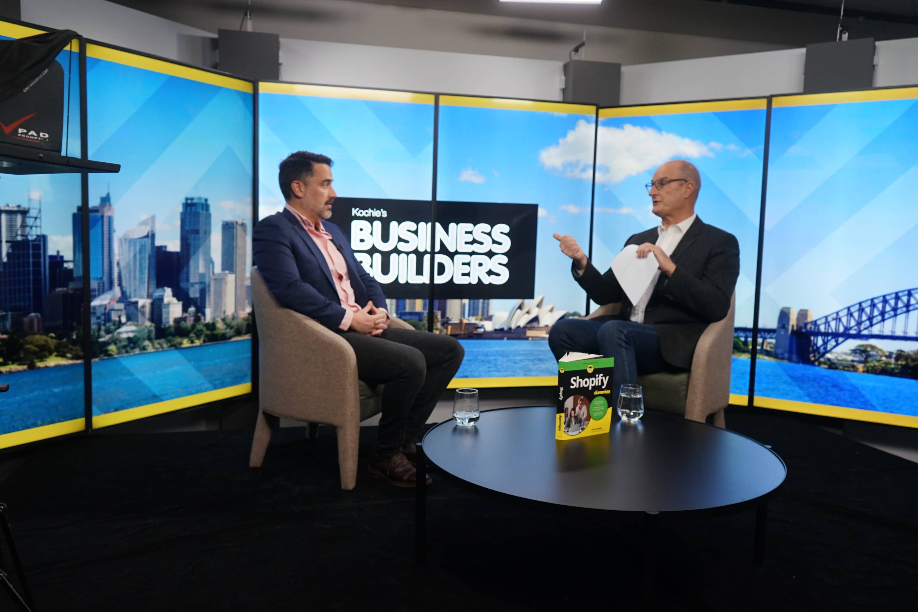A television studio setting features two men seated in chairs, discussing in front of a backdrop of a city skyline, with a promotional book for Shopify on the table.