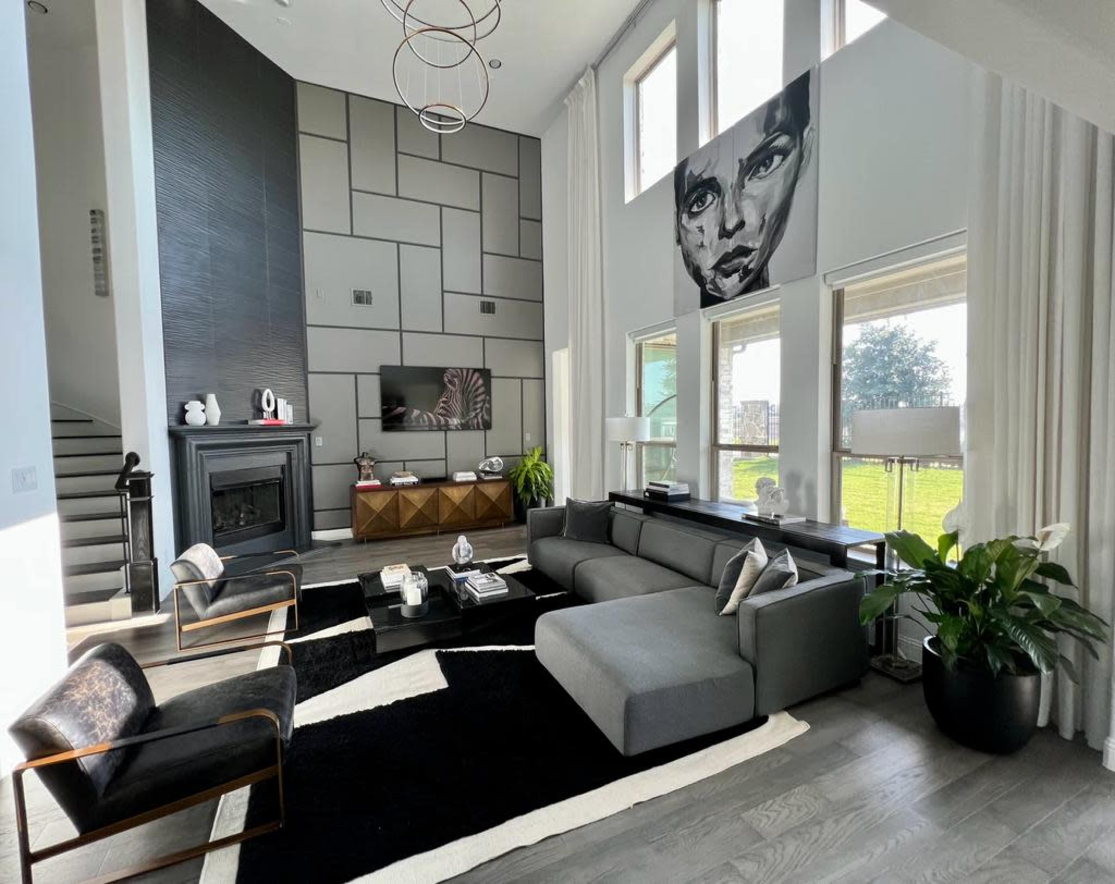 A modern living room with a large gray sectional sofa, black accent chairs, a coffee table, and a prominent wall art piece above a TV and fireplace, surrounded by large windows.