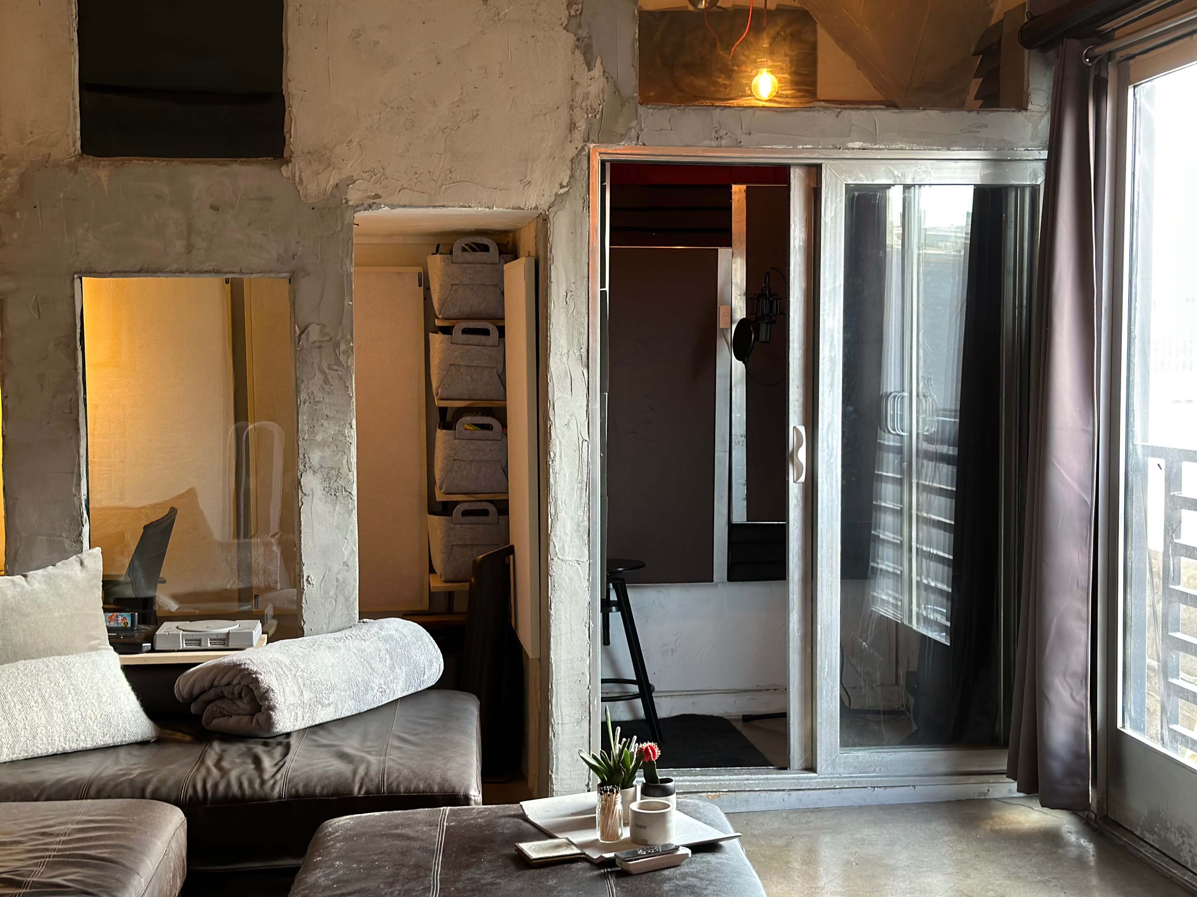 A cozy living area with a sofa, a small table, and a glass door leading to a balcony, alongside a storage area with shelves.