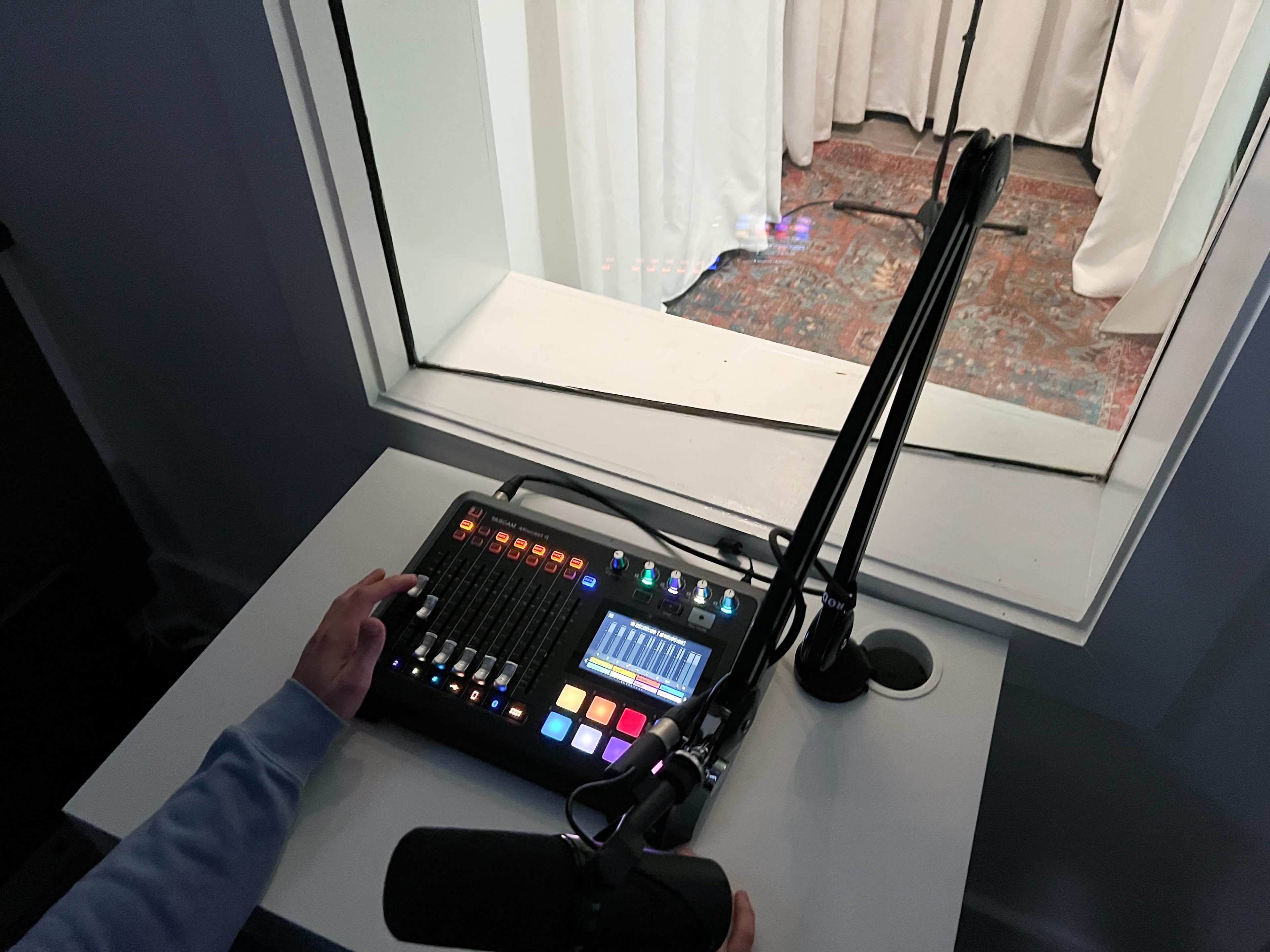 A person operates a sound mixing board with colorful buttons and controls, positioned next to a microphone, while looking through a glass window at a recording area.