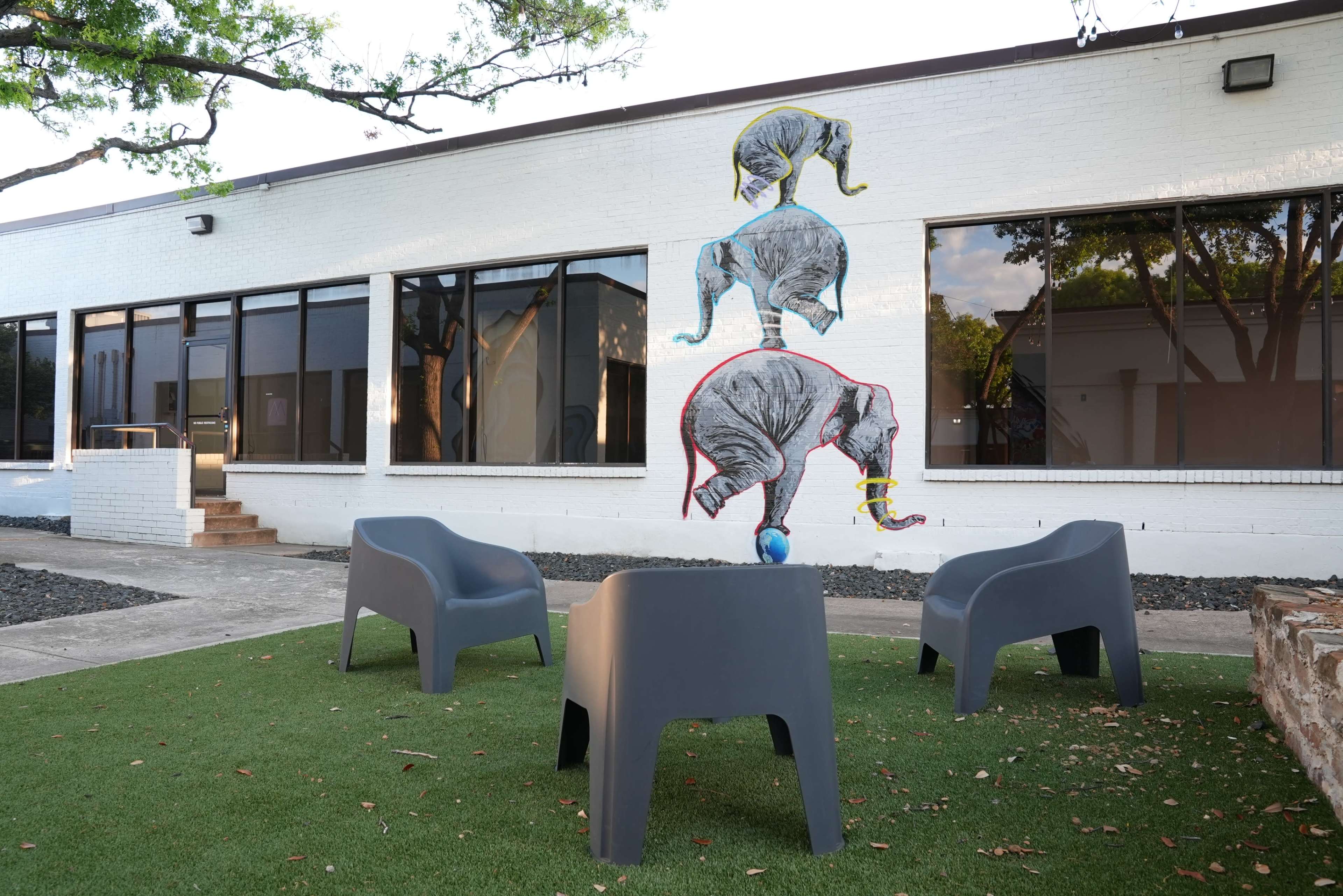The image shows a mural of three elephants arranged in a stack on the white wall of a building, with a seating area consisting of gray chairs on a grassy surface in front.