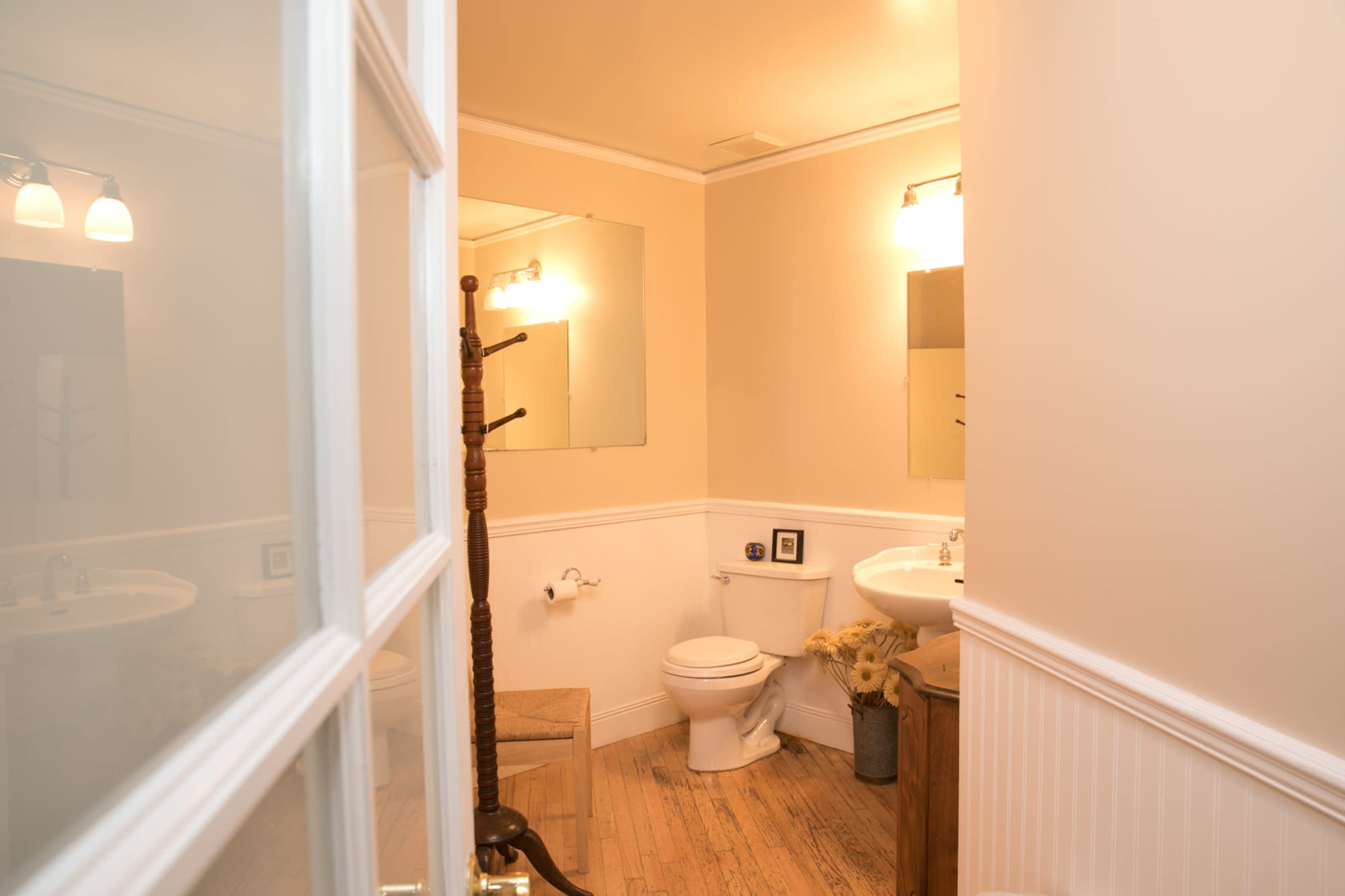 A small bathroom features a toilet, sink, and mirror, with a wooden chair and coat rack near the door.