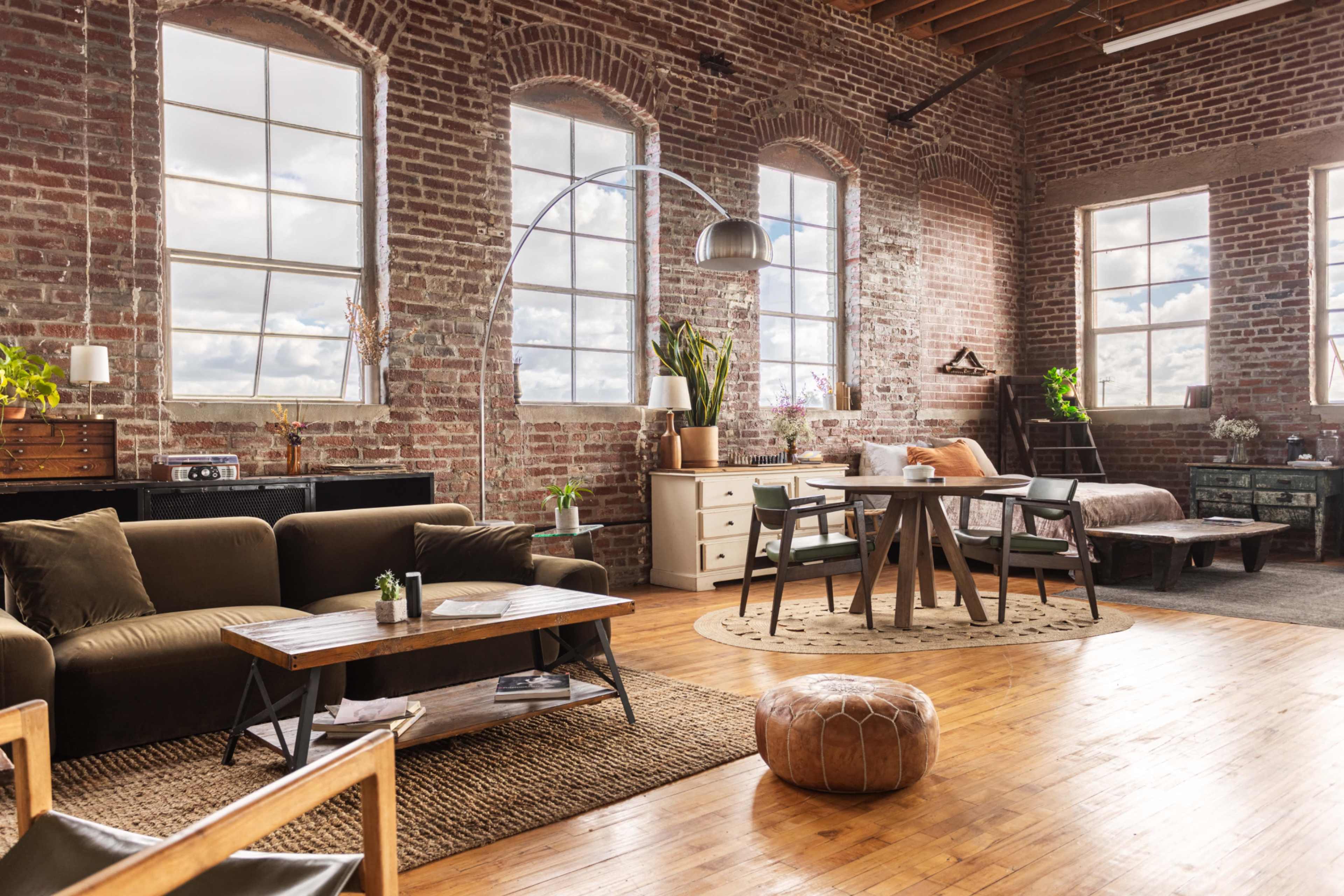 A spacious, industrial-style loft with exposed brick walls, large windows, and a mix of modern and vintage furnishings, including a couch, dining table, and decorative plants.