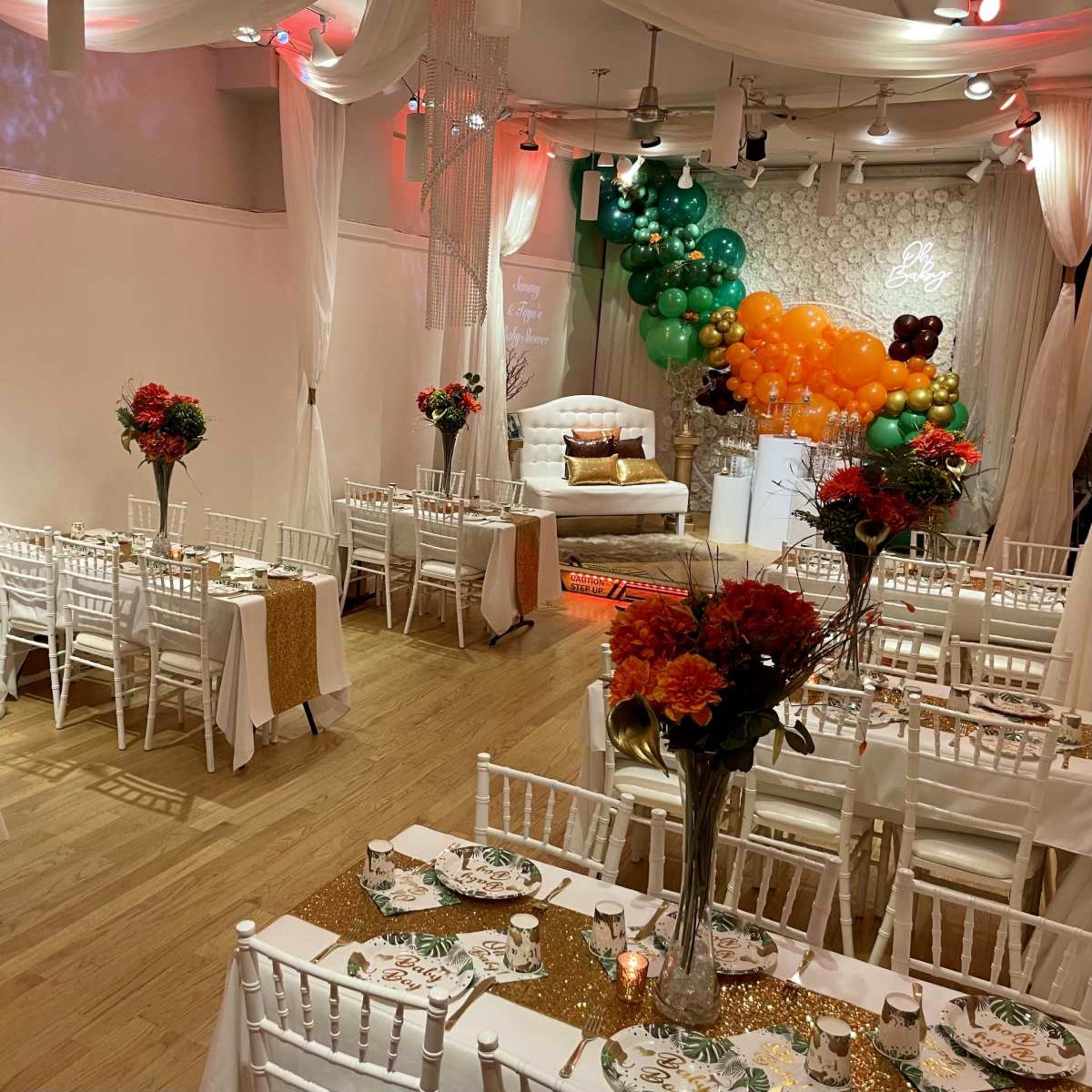 A decorated event space features tables set with floral centerpieces, elegant tableware, and a backdrop of balloons and drapery, creating an inviting atmosphere for a celebration.