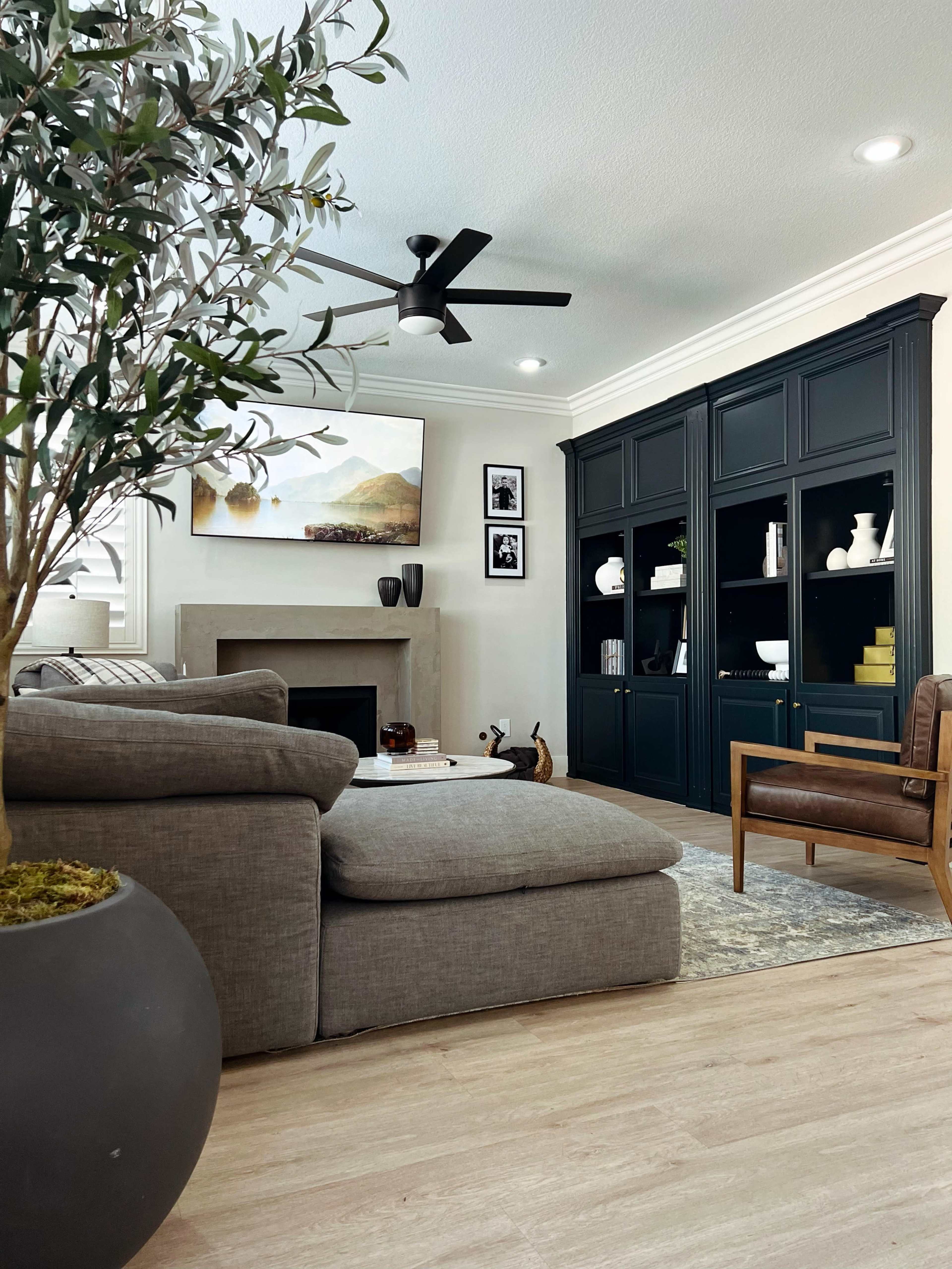 A cozy living room features a large sectional sofa, a modern ceiling fan, a fireplace, and built-in shelves filled with decorative items.