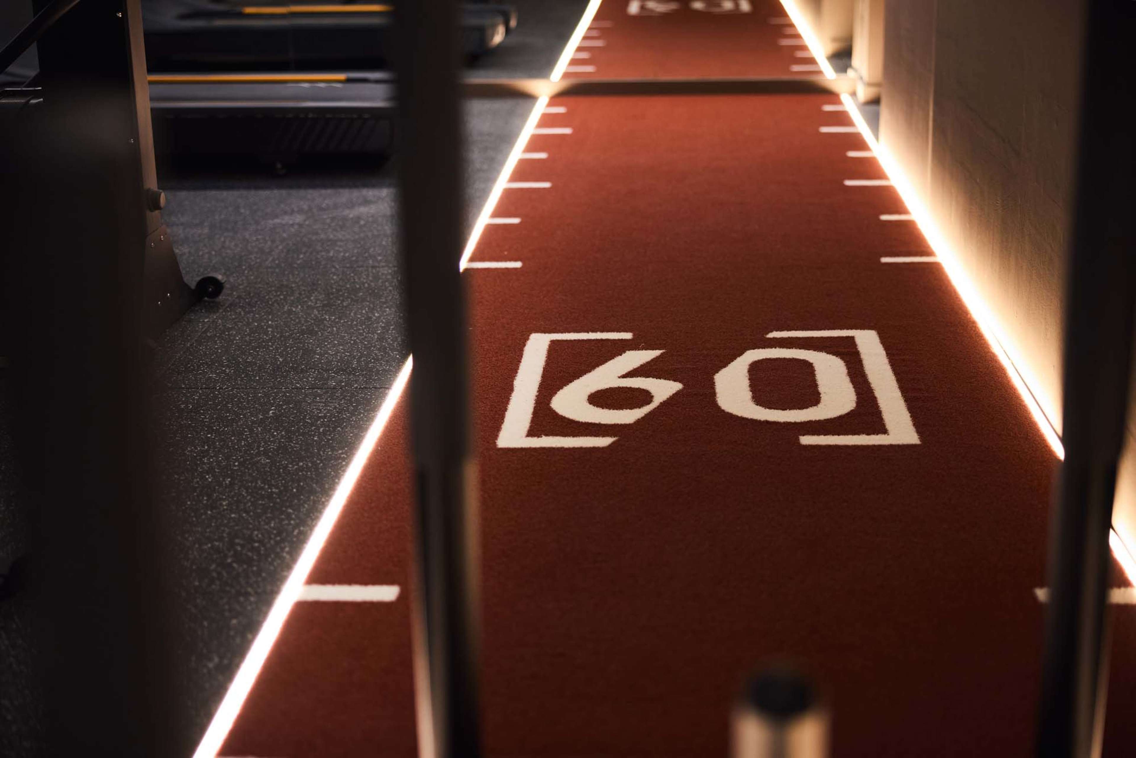The image shows a red carpet with a white number "60" marked on it, situated in a gym or exercise area with illuminated edges.