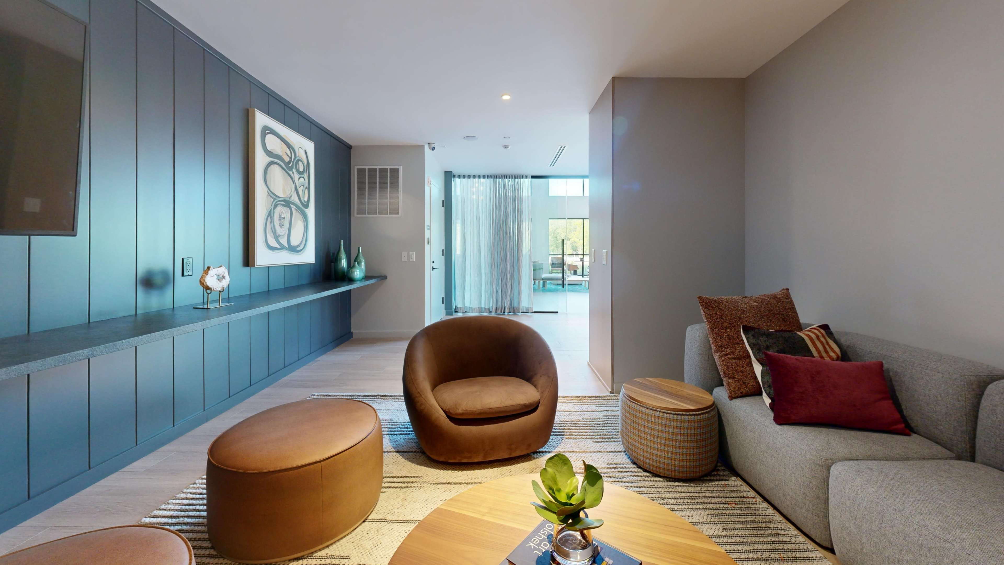 The image shows a modern living room featuring a brown armchair, a gray sofa, a round coffee table, and a textured area rug, with a large abstract painting on the wall and glass doors leading to an outdoor space.