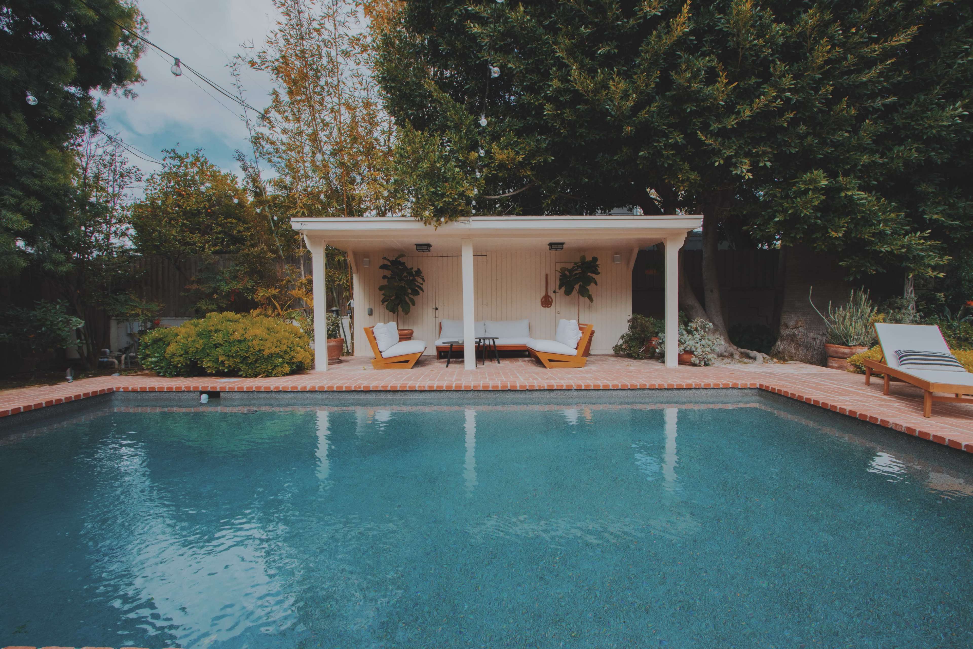 A pool with clear water is situated in front of a covered seating area surrounded by greenery and potted plants.