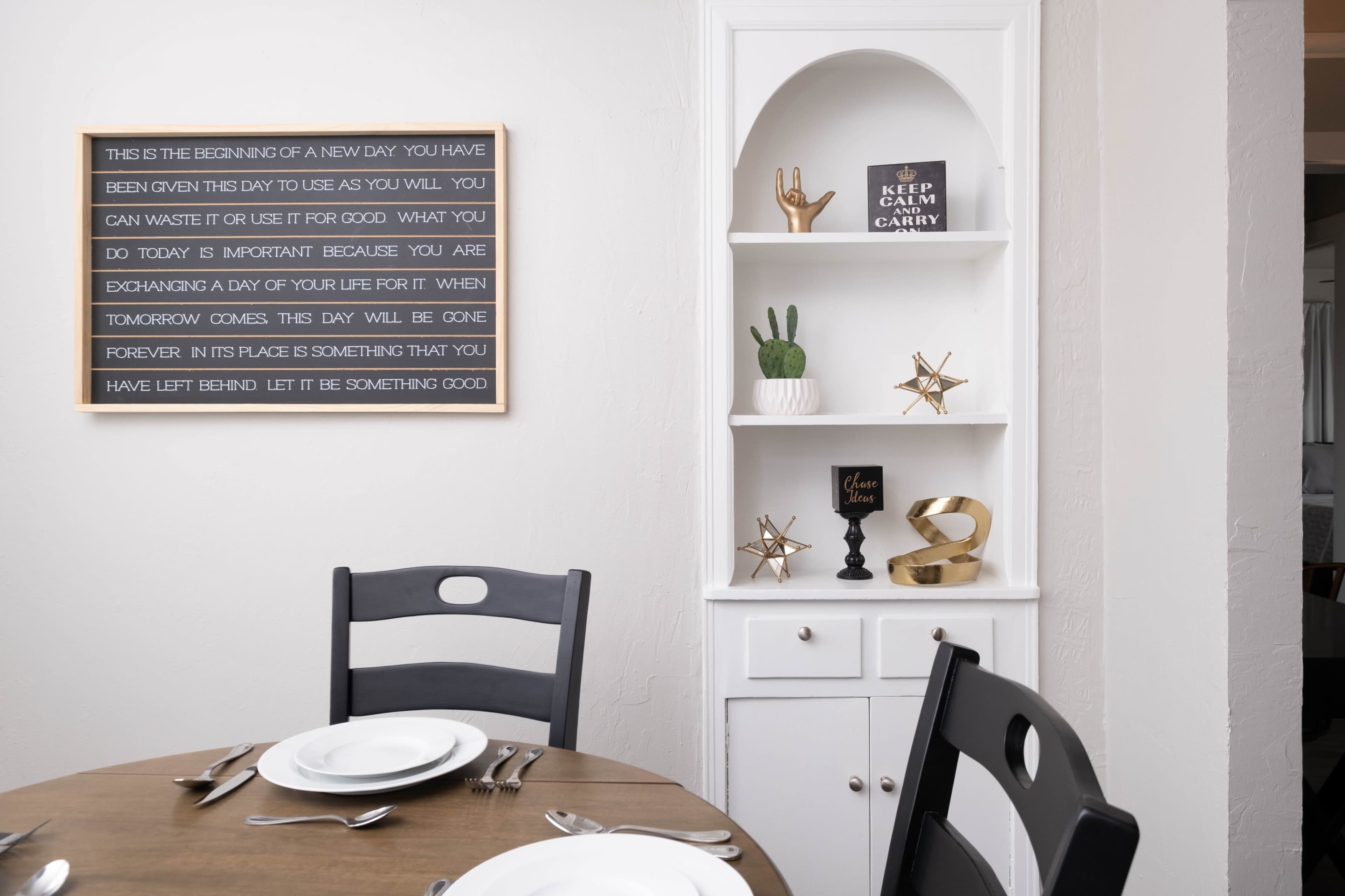 A table is set for a meal in a dining room featuring a wall with inspirational quotes and a white shelf displaying decorative items.