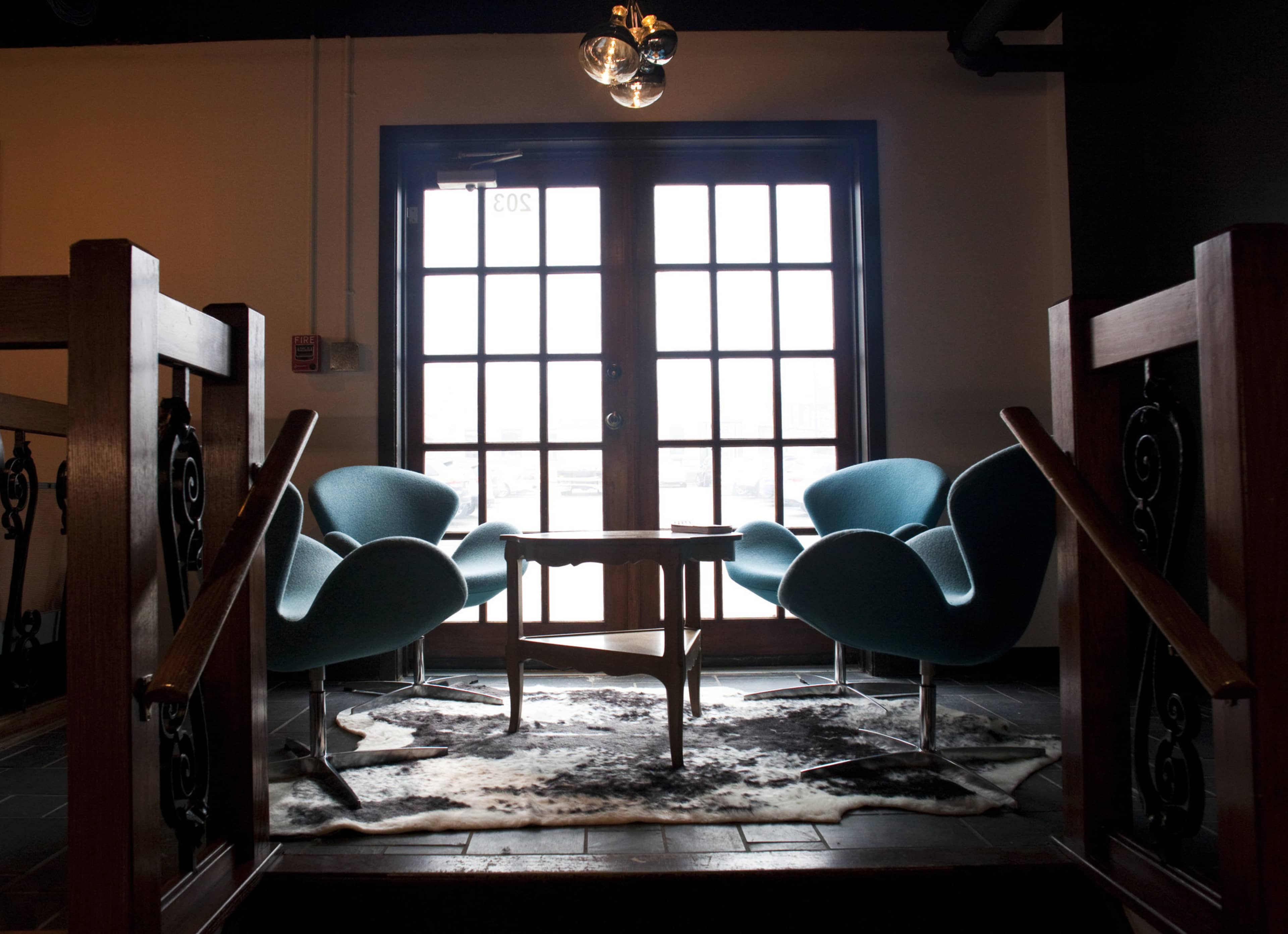 A pair of teal chairs and a wooden table are positioned near a large window and set on a cowhide rug at the top of a staircase.