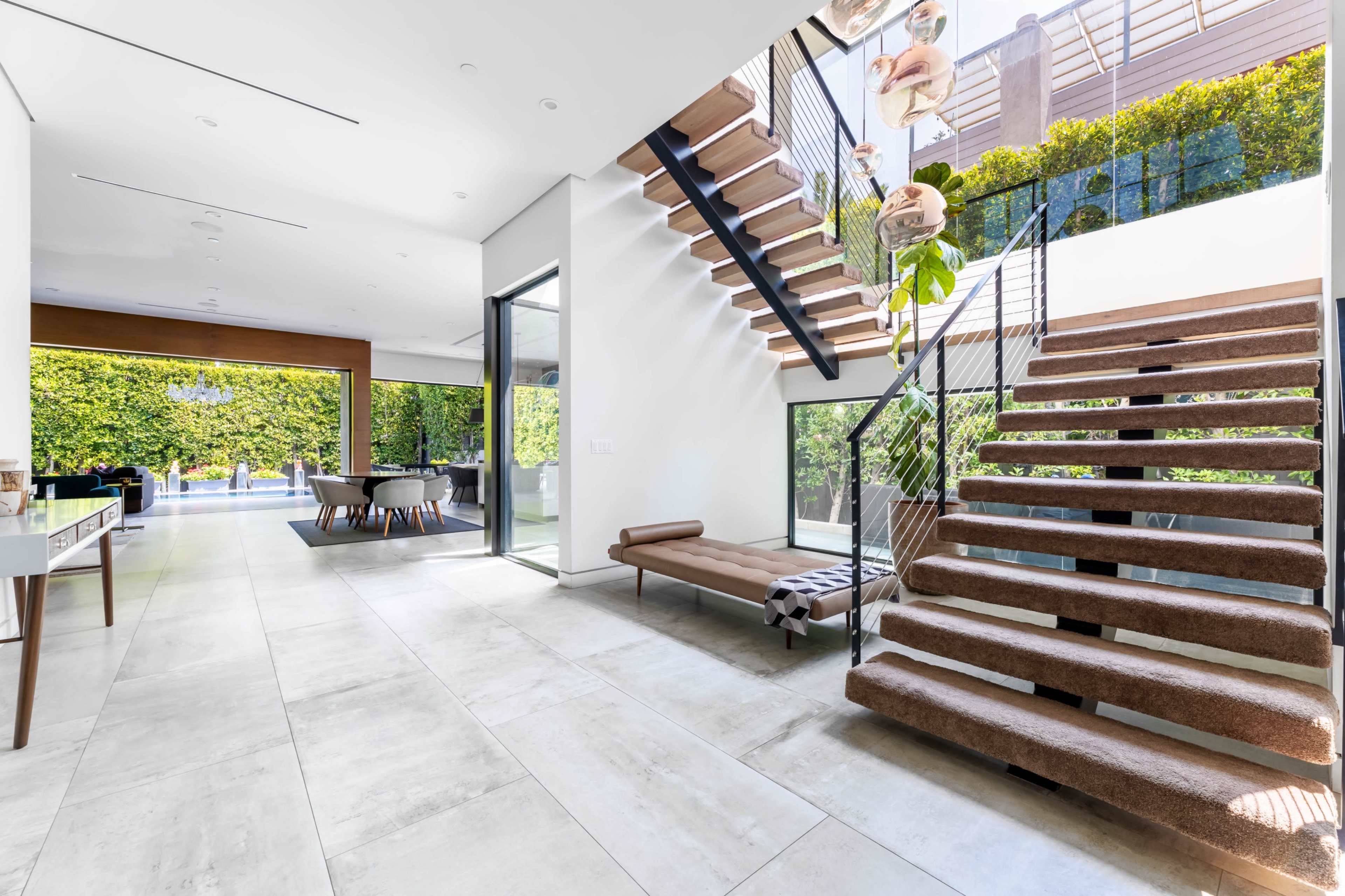The image shows a modern interior space featuring a staircase with a glass railing, large windows, and a view of an outdoor area with greenery.
